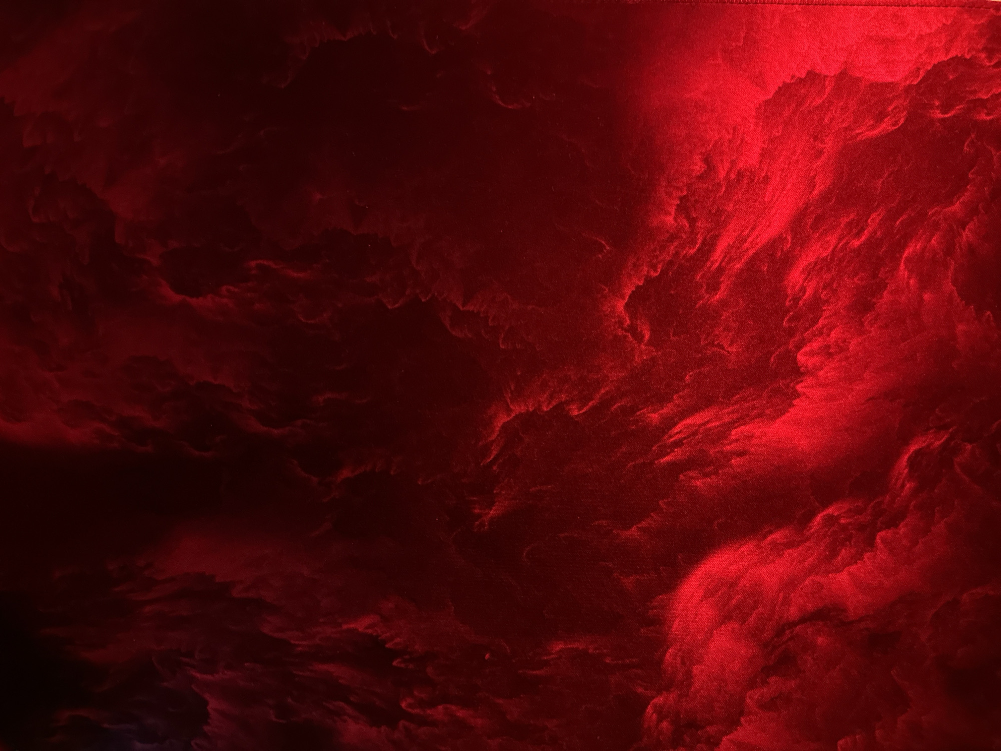 A close-up, overhead, low-angle shot shows a dark red, textured surface with swirling patterns that resemble clouds or smoke. The lighting is dramatic, with bright red light illuminating some areas and casting others into deep shadow. The texture appears to be slightly granular. The image is abstract and does not depict any recognizable objects or environments.
