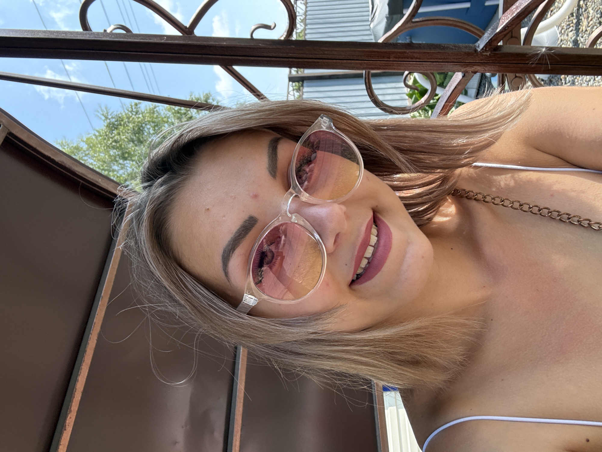 A young woman with blonde hair and sunglasses is looking at the camera outdoors. She is wearing a white strap top. The background shows a fence, trees, and a cloudy blue sky. The image appears to have been taken in Krasnogorskoye, Russia.