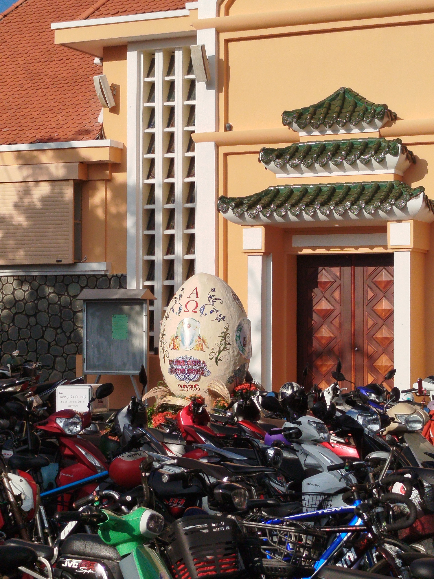 The image presents a street view in Ho Chi Minh City, Vietnam, dominated by a multi-tiered building with an orange facade and a tiled roof. A large, decorated egg-shaped monument stands prominently near the entrance, adorned with religious symbols and the inscription "2026". The entrance features a dark wooden double door beneath a series of ornate, layered green tiled eaves. The building also has a distinct window design with white vertical and horizontal elements. In front of the building, a dense collection of motorbikes is parked, obscuring part of the view. A small sign stands near the motorbikes, bearing the text "GÓP QUỸ CỨU TRỢ" followed by Vietnamese text. The overall impression is one of a busy urban scene with religious and cultural elements.