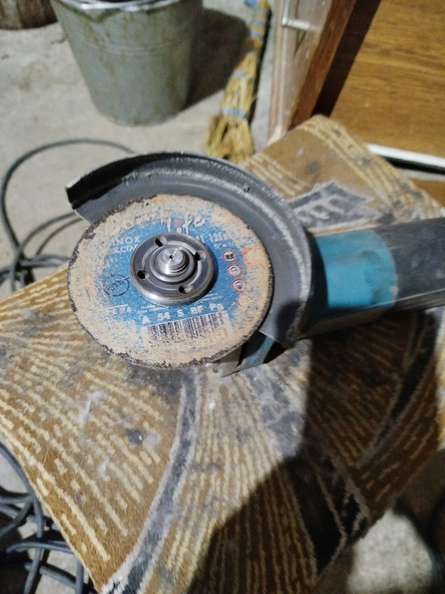 The image shows a close-up of a used angle grinder resting on a patterned rug. The angle grinder has a blue and white cutting disc attached, which appears dusty and worn. The grinder itself is primarily black and teal. In the background, a metal bucket, a broom with a straw head, and a wooden structure are partially visible. The setting appears to be indoors, possibly a workshop or storage area, within Крывасельскі сельскі Савет, Belarus. There are no people or animals in the frame. The overall scene suggests a functional, perhaps industrial or DIY, environment.