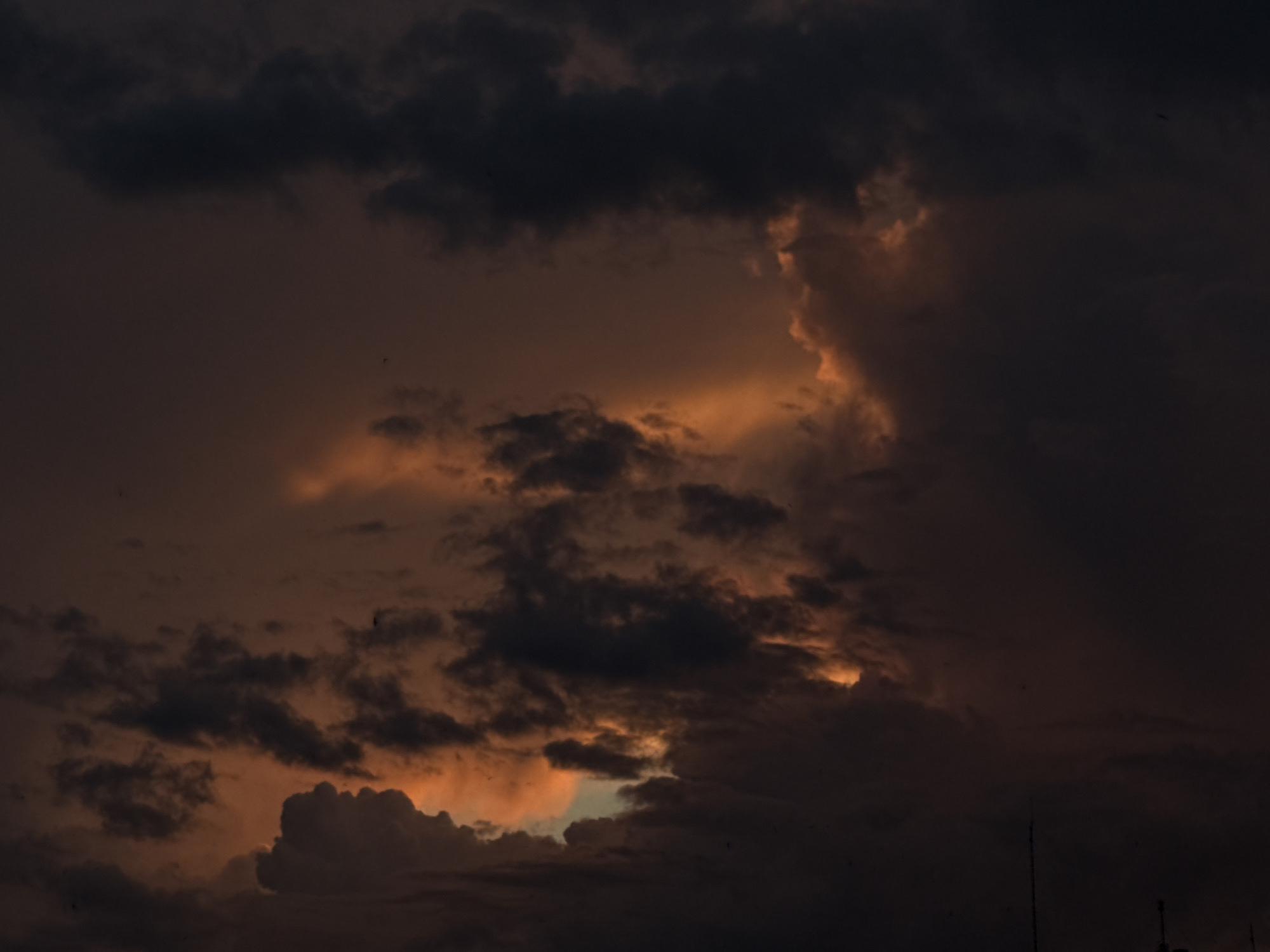 A low-angle, wide shot captures a dramatic sky over the city of Luhansk, Ukraine. The scene is dominated by dark, ominous clouds tinged with shades of orange and red, suggesting either a sunset or a storm approaching. The lighting is dim, with pockets of brighter, fiery color peeking through the dense cloud cover. The overall mood is somber and foreboding. There are no visible people or specific landmarks to indicate the precise location within Luhansk, but the presence of what appear to be distant structures at the very bottom of the frame suggests an urban or semi-urban environment.