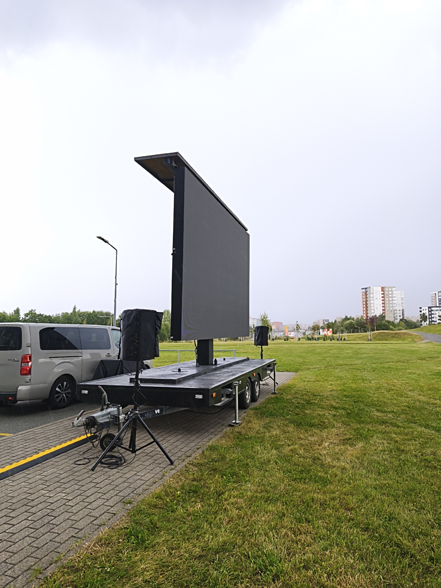 In Tallinn, Estonia, on a cloudy day, a large mobile LED screen is set up on a trailer in a grassy area adjacent to a paved walkway. A light-colored van is parked on the walkway to the left of the trailer. The LED screen is large and black, with a protective canopy angled upwards. Two black speakers are visible on the trailer, one in front of the screen and another to the left. A tripod with cables is positioned on the paved area in front of the trailer. In the background, there is a row of trees and several multi-story buildings, suggesting an urban or suburban environment. The ground is covered with green grass and the sky is overcast.