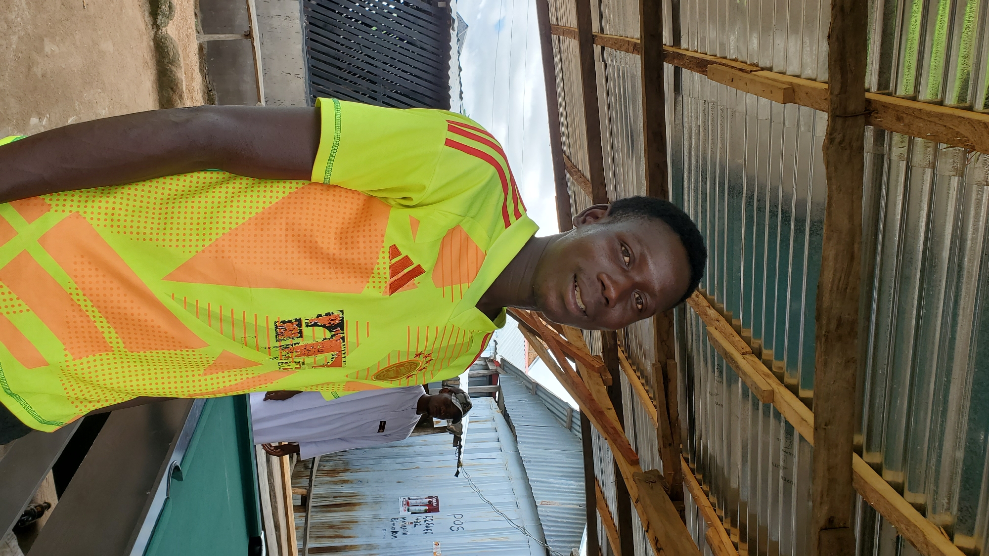 A young man wearing a bright yellow and orange athletic shirt with red accents stands looking at the camera. He is in the foreground of the image, with his head and torso visible. Behind him, another man in a white robe and a cap is partially visible near a green pool table. The scene appears to be outdoors, under a corrugated metal roof supported by wooden beams, suggesting a covered outdoor area. The environment includes a wooden fence and what looks like a building in the background with some signage. This setting is consistent with informal gathering places found in Jalingo, Nigeria, where outdoor recreational areas are common.