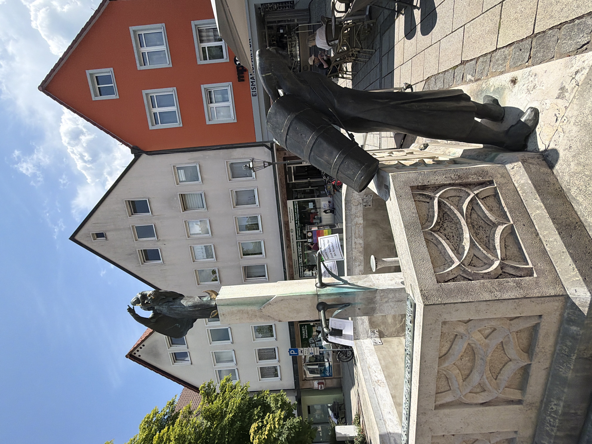 The image depicts a public square in Ebermannstadt, Germany, featuring a bronze sculpture of a person carrying a large barrel. The sculpture is located in front of a fountain with a decorative stone basin. In the background, there are several buildings, including a prominent orange-colored structure and a light-colored, multi-story building with numerous windows. A lamppost stands near the orange building. An awning is extended over a cafe or restaurant area with outdoor seating, where two people are visible sitting at a table. The ground is paved with cobblestones. A tree with green foliage is visible on the left side of the frame. The sky is blue with scattered white clouds. The scene appears to be from a sunny day.