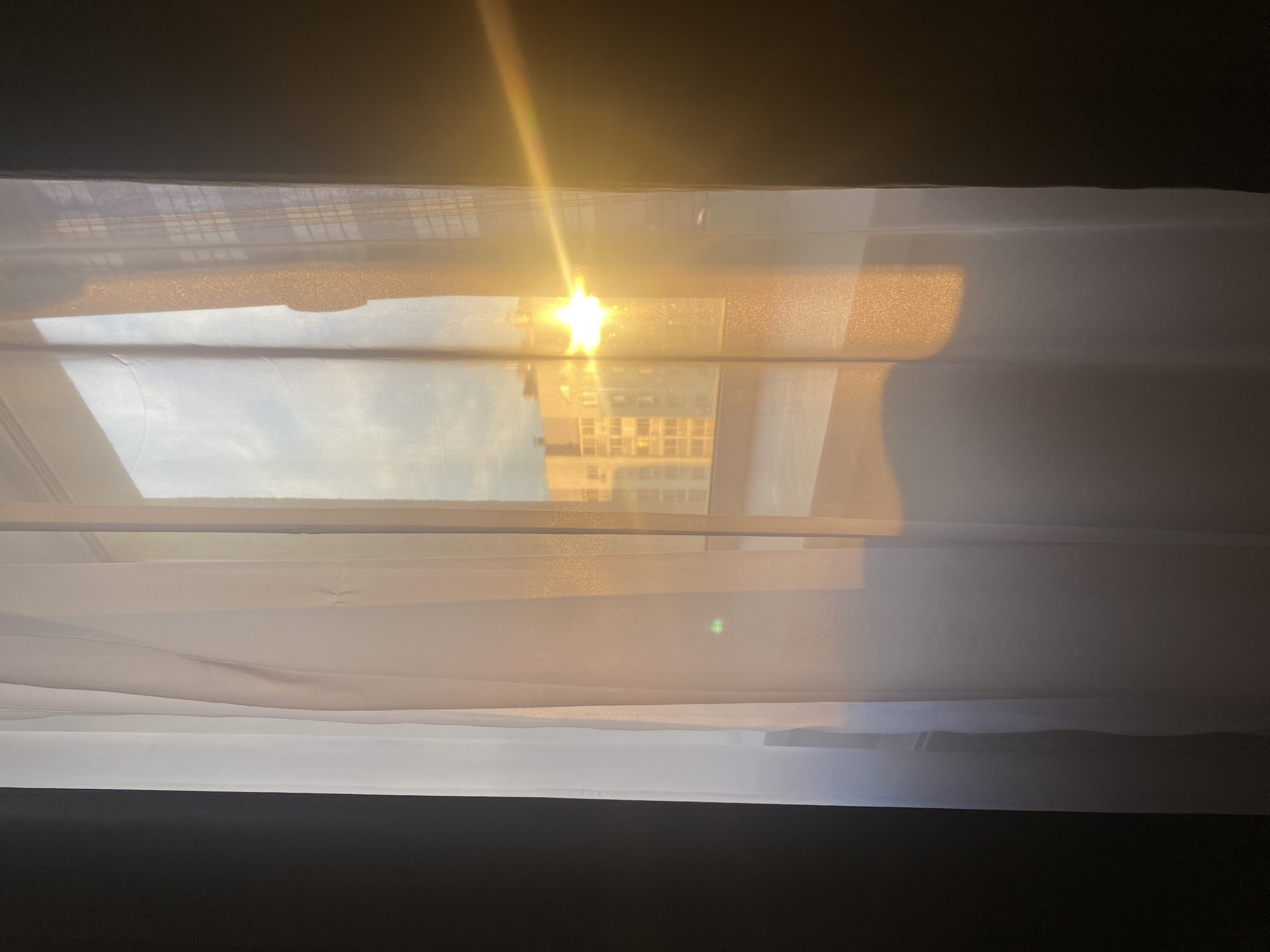This media file captures a view through a window, likely from inside a building, looking out at a city scene during sunset. The foreground is dominated by sheer white curtains, which are partially drawn and create soft folds and shadows. The curtains diffuse the light, giving a hazy, ethereal quality to the image.
Beyond the curtains, a building with multiple floors and windows is visible, reflecting the warm glow of the setting sun. The sun itself is a bright, starburst-like light source near the top of the frame, casting a strong glare and creating a lens flare. The sky appears to be a pale blue with some hints of clouds. The overall atmosphere suggests a peaceful and quiet moment, possibly the end of a day.
The light suggests it is late afternoon or early evening, as indicated by the golden hues and the low angle of the sun. The presence of a city building implies an urban or suburban setting. There is no visible text in the image. The scene evokes a sense of calm and perhaps a touch of melancholy associated with the end of daylight.