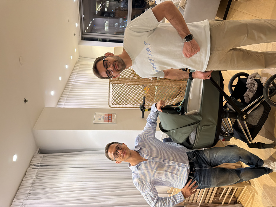 Two men wearing glasses pose in a modern living room. One wears a white t-shirt and beige pants, while the other wears a blue striped shirt and jeans. A baby stroller is between them. The room features light-colored walls, white curtains, a decorative screen, and a "MAKE ART NOT WAR" poster. A night view is visible through a large window.