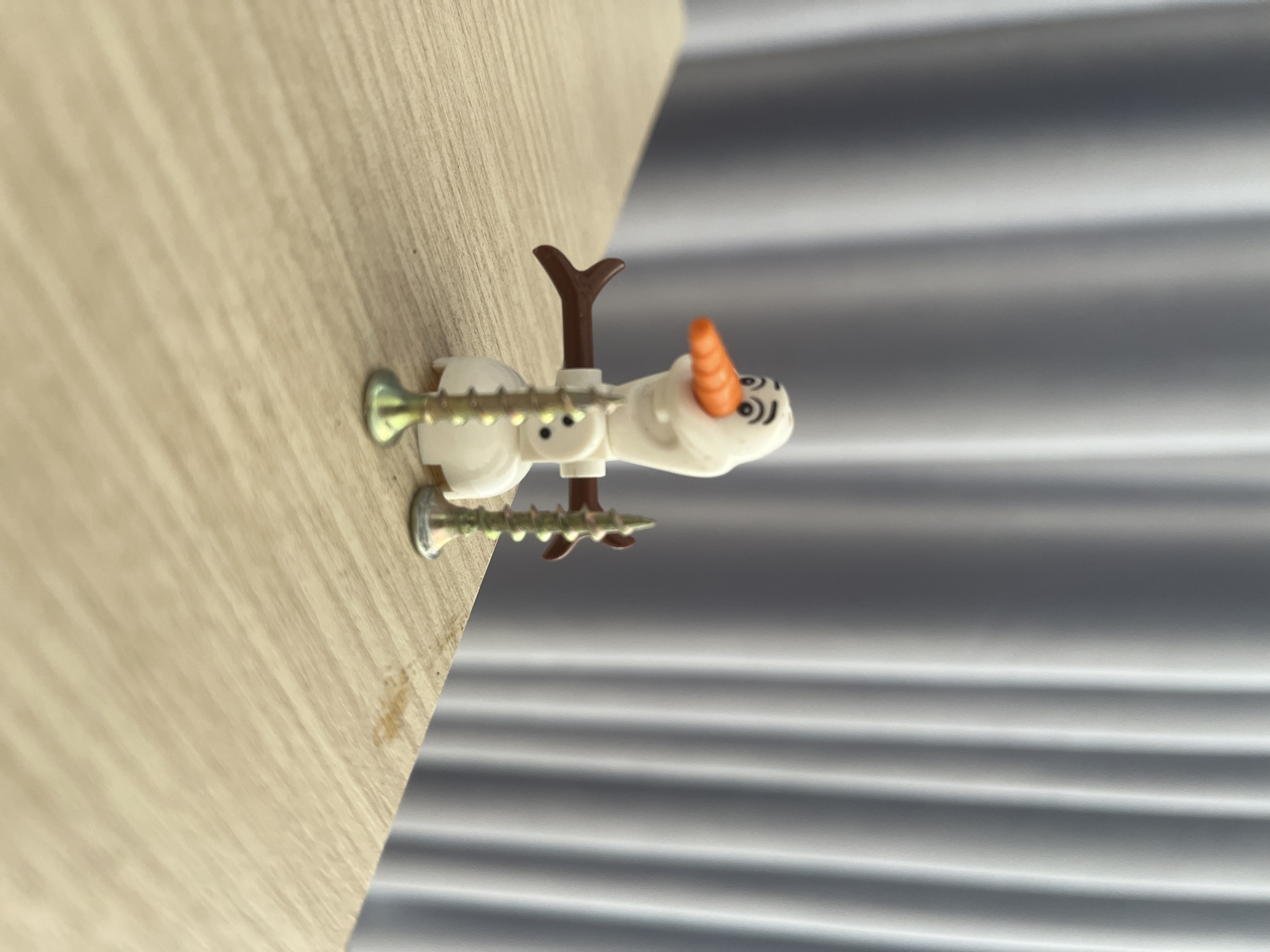 A sideways close-up shows a plastic Olaf figurine impaled by two screws and affixed to a light-colored wooden surface. The background consists of horizontally oriented, soft, silver-grey drapery with linear patterns. There is no visible indication of Bac Nha Trang Ward, Vietnam.