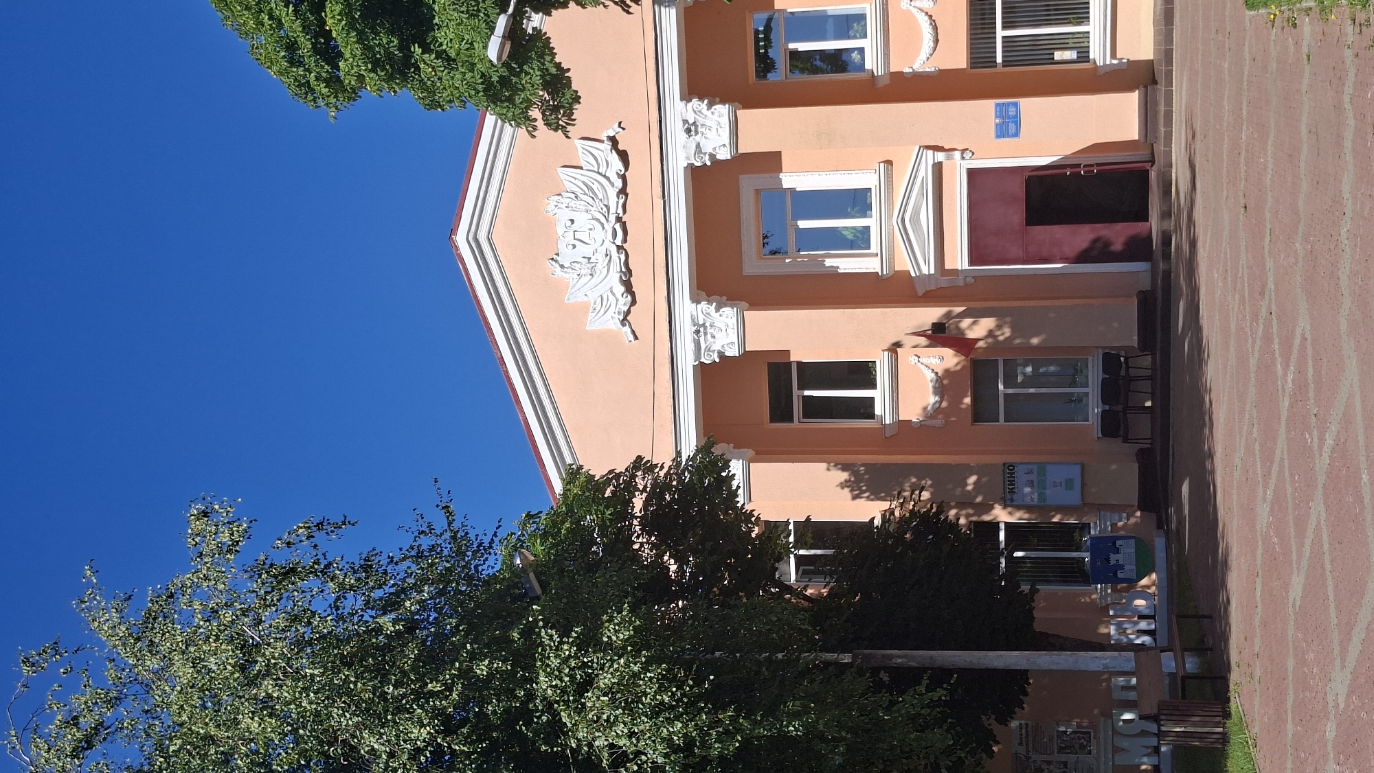 The image shows the facade of a building in the city of Myadzyel, Belarus, which appears to be a cultural center or cinema based on the signage. The building has a salmon-pink color with white decorative elements, including columns and a relief on the pediment. Several windows with white frames are visible, as is a large dark red entrance door. To the left of the entrance, there is a sign with "КИНО" (KINO) written on it, indicating a cinema. A red flag hangs on the right side of one of the windows. In front of the building, there is a paved area with a brick pattern. Trees with green foliage are present on both sides of the building, partially obscuring the facade. A bench and a trash can are also visible in the foreground to the left. The sky is clear and blue.