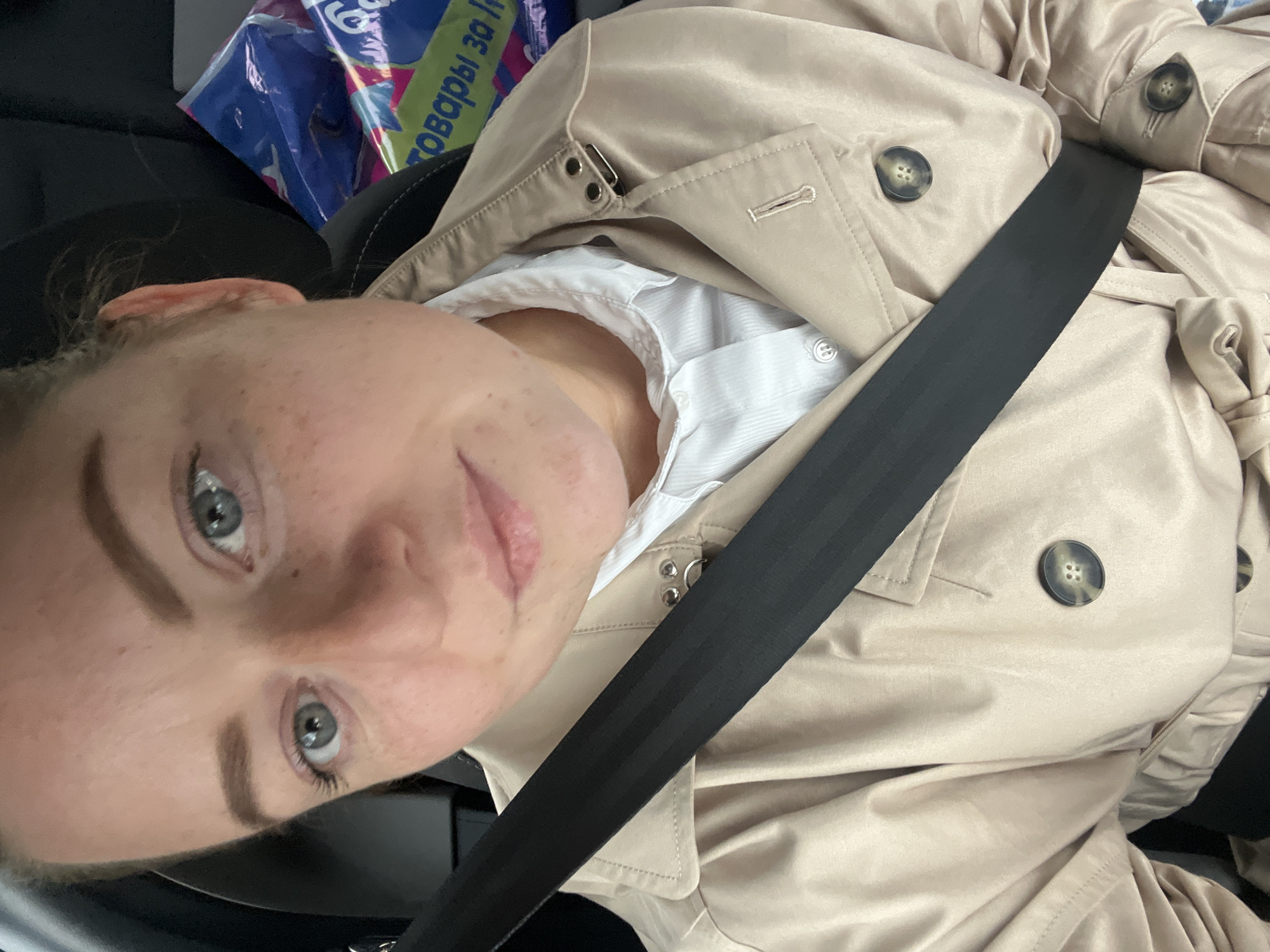 A medium shot from a tilted angle features a fair-skinned woman with freckles and light eyes, wearing a white shirt and a beige trench coat, secured by a black seatbelt. The setting appears to be inside a vehicle. In the upper left corner, a blurred object with blue, purple, and green packaging is visible. The text on the package includes "товары за 30" (goods for 30). The location in the city of Ширяево, Russia, cannot be verified from the information provided in the picture.