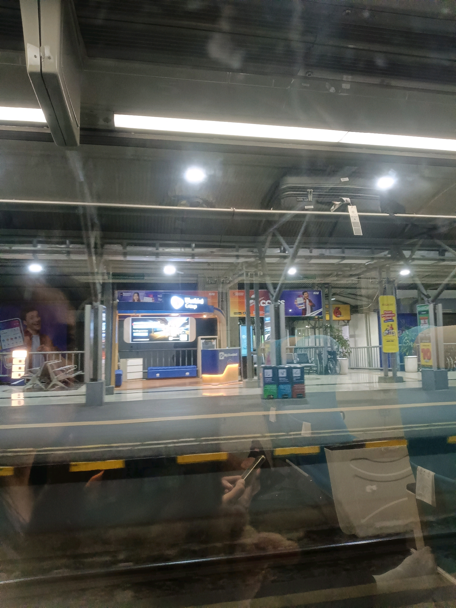 This image shows a view from inside a train car at a station in Surabaya, Indonesia. The station appears to be an elevated platform with a roof structure supported by metal beams. Fluorescent lights are visible along the ceiling.
On the platform, there are several kiosks and advertisements. One kiosk has a blue and white design with screens displaying images. Another kiosk features a large advertisement with a picture of a man and text. There are also some empty chairs and a bicycle visible. Several trash bins are lined up near a kiosk.
Reflections on the train window show parts of the interior of the train, including the hands of passengers holding smartphones. The platform is separated from the train tracks by a railing and a dark strip with yellow markings. The overall scene suggests a busy urban transportation hub.