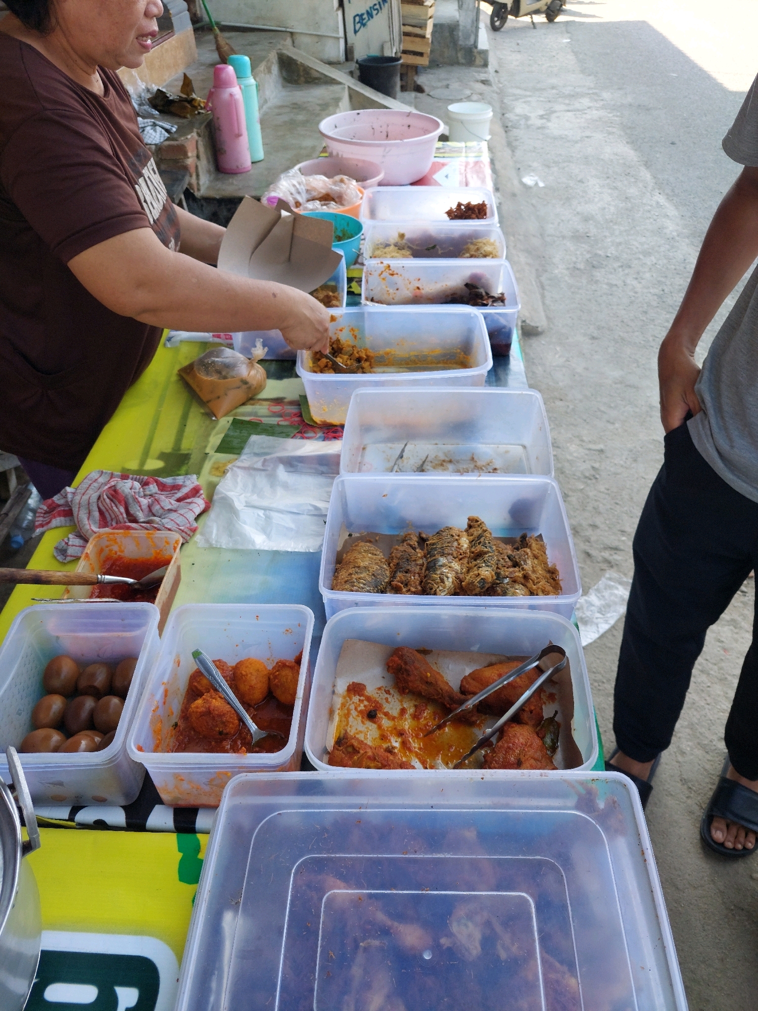 This is a medium shot of a street food stall in Arjowilangun, Indonesia. A woman in a brown t-shirt is serving food from several clear plastic containers on a yellow-and-green patterned table cloth. She is holding a brown paper container and a spoon in her hands. In front of her are various food items in plastic containers, including what appears to be boiled eggs, fried fish, and some kind of stewed dish with meatballs. There are also some other containers and a pink and blue thermos bottle visible. To the right of the woman, a person wearing black pants and black sandals stands with their hands in their pockets. The background shows a paved street and a building. The overall scene depicts a typical street food vending operation in Indonesia.