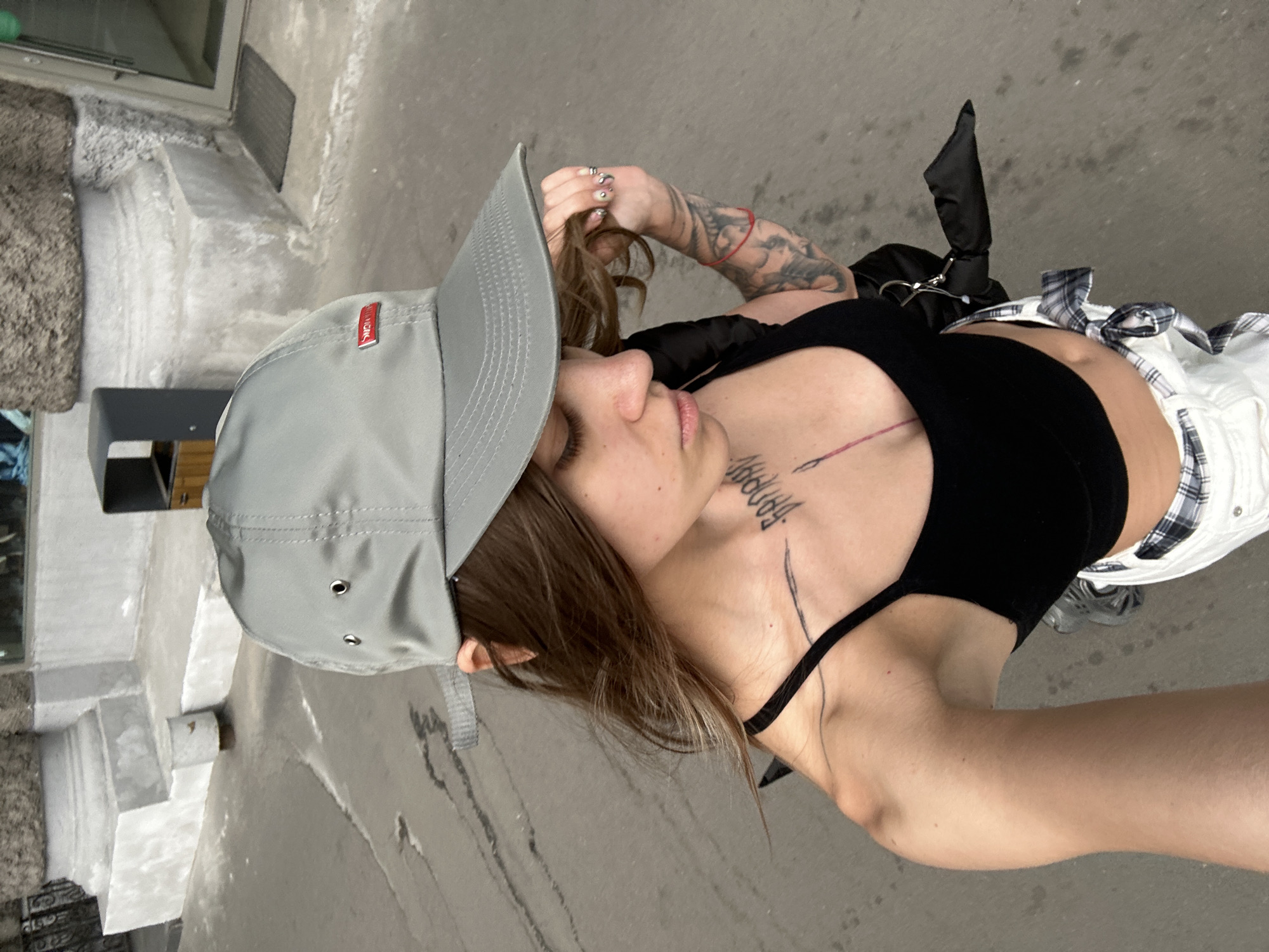 An outdoor, eye-level, medium shot shows a young, white woman from the shoulders up as she takes a selfie outdoors in a city. She is wearing a light gray baseball cap, a black spaghetti strap crop top, and white pants with a plaid pattern visible at the waist. Her hair is long and brown, falling over her shoulders. She has tattoos on her left arm and chest. The background consists of a sidewalk and a building with a large stone column. The overall lighting is natural and bright, suggesting it is daytime. The specific location is identified as Moscow, Russia.