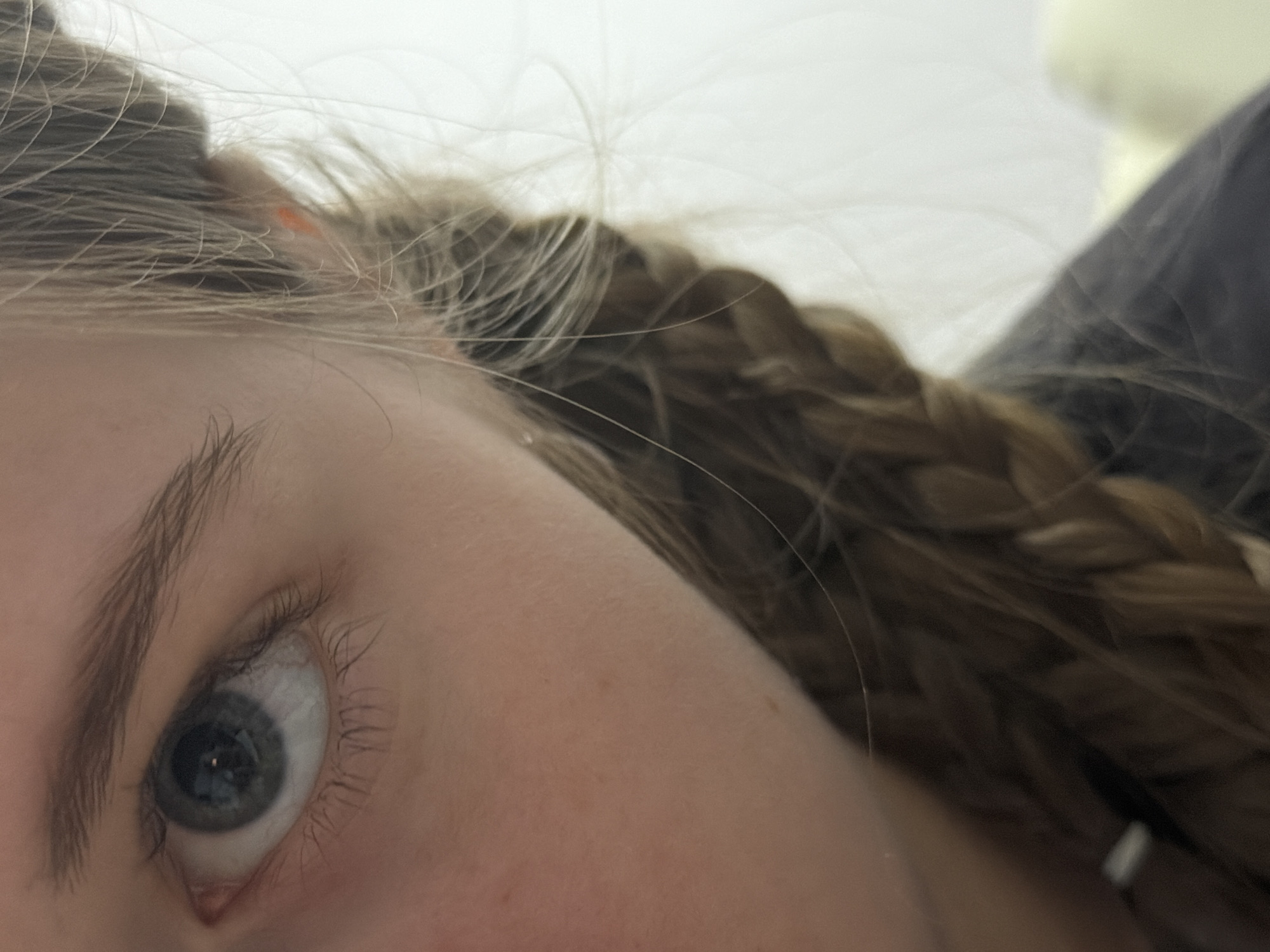 This close-up, diagonally composed image focuses on the left eye and surrounding features of a person, likely female. The dominant subject is a light blue or grey eye, with a clearly visible pupil reflecting a light source. Long, natural eyelashes frame the eye, and a well-defined eyebrow arches above it. The skin, appearing fair, shows subtle texture and perhaps a few minor freckles on the cheek area.
Light brown or blonde hair, mostly styled in a braid, runs from the top center towards the upper right of the frame. Numerous individual strands of hair are visible against the blurred background, some straying across the lighter areas. A small, orange accessory, possibly a hair tie, is faintly discernible within the hair near the upper left.
The background is largely out of focus. The upper and left portions show a light, uniform color, potentially a wall or soft bedding, while the right side features a darker, indistinct shape that could be part of a pillow or blanket. The lighting is soft and even, suggesting an indoor setting without harsh shadows. The person's gaze appears direct and focused, looking slightly upwards and to their left. There is no visible text in the image.