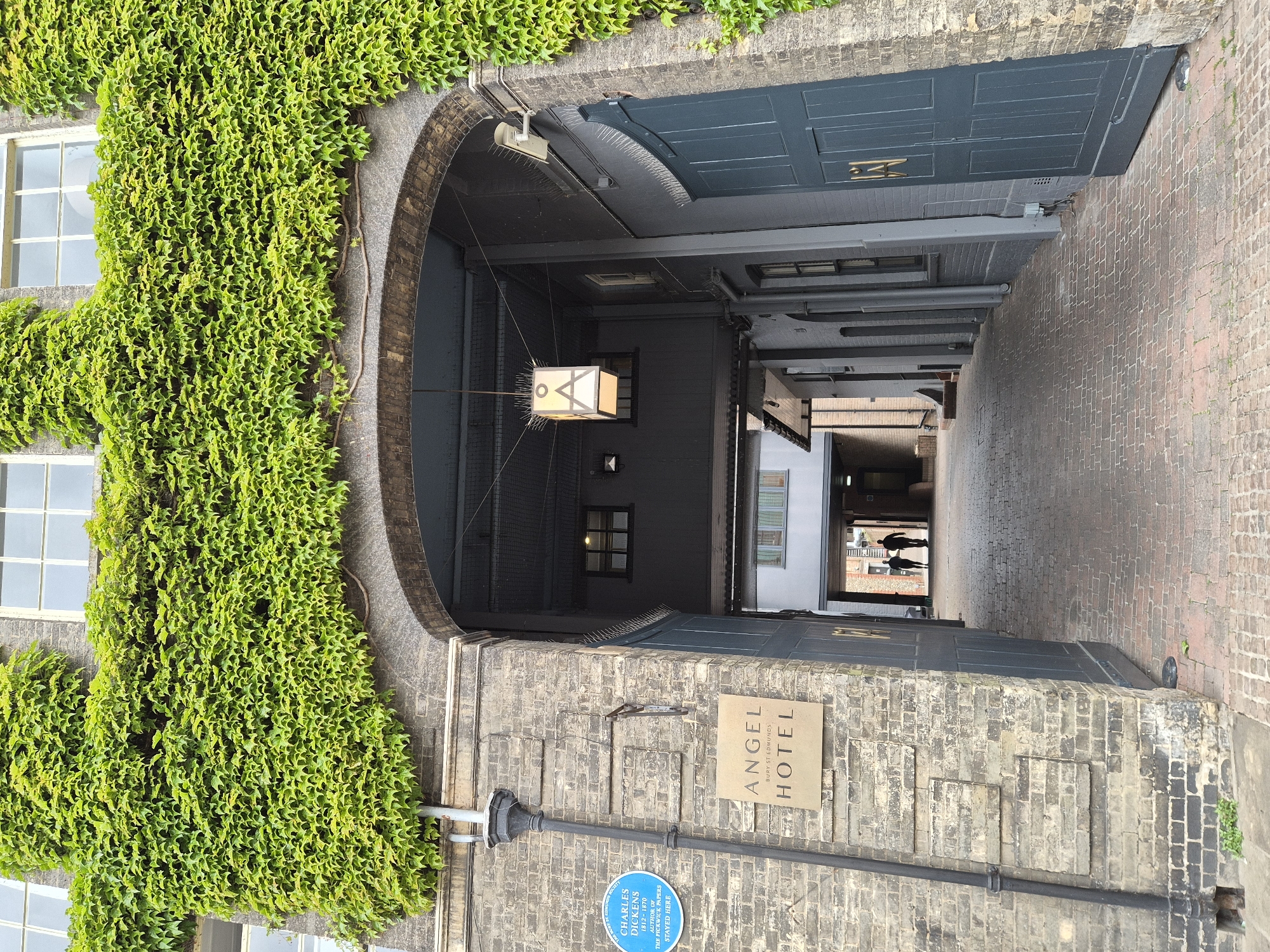 The image shows an alleyway leading through a brick archway in Bury St Edmunds, West Suffolk, UK. The left side of the entrance is covered in dense green ivy. To the right, the facade of a brick building is visible, with a sign reading "ANGEL BURY ST EDMUNDS HOTEL" mounted on the wall. Below the hotel sign is a blue plaque identifying Charles Dickens as having stayed there. The alleyway is paved with brick and lined with buildings, diminishing in depth towards the exit where two figures are walking away from the camera. An ornate light fixture hangs from the ceiling of the archway.