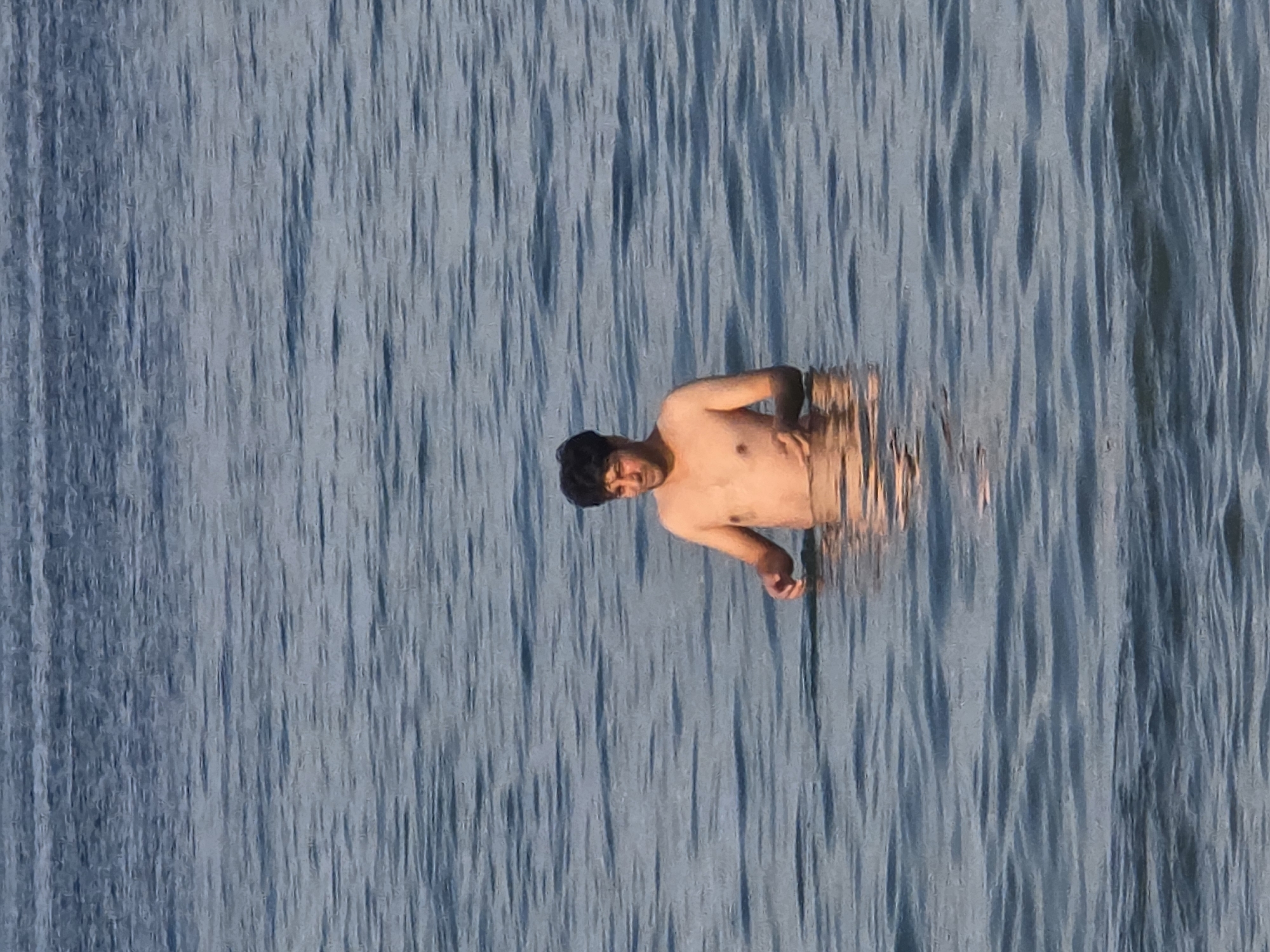 A man is standing waist-deep in rippling, blue water, facing the camera with a slight smile. His skin is sun-kissed, and he has dark, curly hair. His hands are clasped in front of him. The water appears calm with gentle waves. The lighting suggests it is daytime, possibly late afternoon or early evening, given the warm tone of the man's skin and the subtle highlights on the water. The background is entirely composed of the water's surface, with no land or sky visible, implying he is in a large body of water such as the Trave river or the Baltic Sea, near Lübeck, Germany.