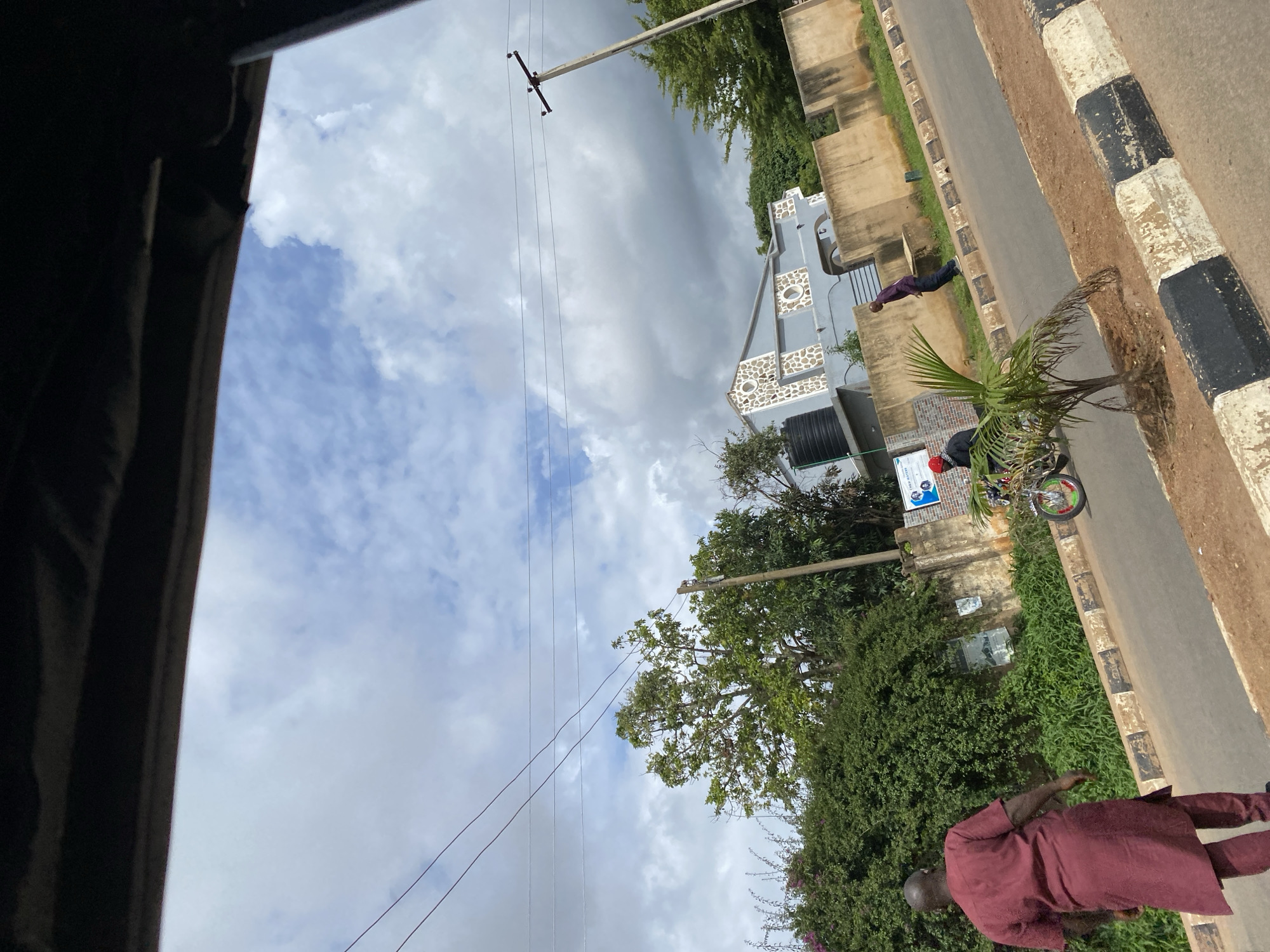 The image captures a view of a street in Bar Jirgi Summa, Nigeria, under a cloudy sky. In the foreground, a man wearing a maroon traditional outfit walks away from the camera along the side of a paved road. The road has a distinct black and white checkered curb. To the left of the road, dense green foliage forms a hedge, behind which a light blue building with a patterned stone facade is visible. A large black water tank is situated near the building. Further down the road, a person on a motorcycle, wearing a red helmet, is seen in motion. Another person in a dark shirt and pants walks away from the motorcycle and the building. A utility pole with wires is present on the right side of the road. The overall scene suggests a residential or commercial area with moderate activity.