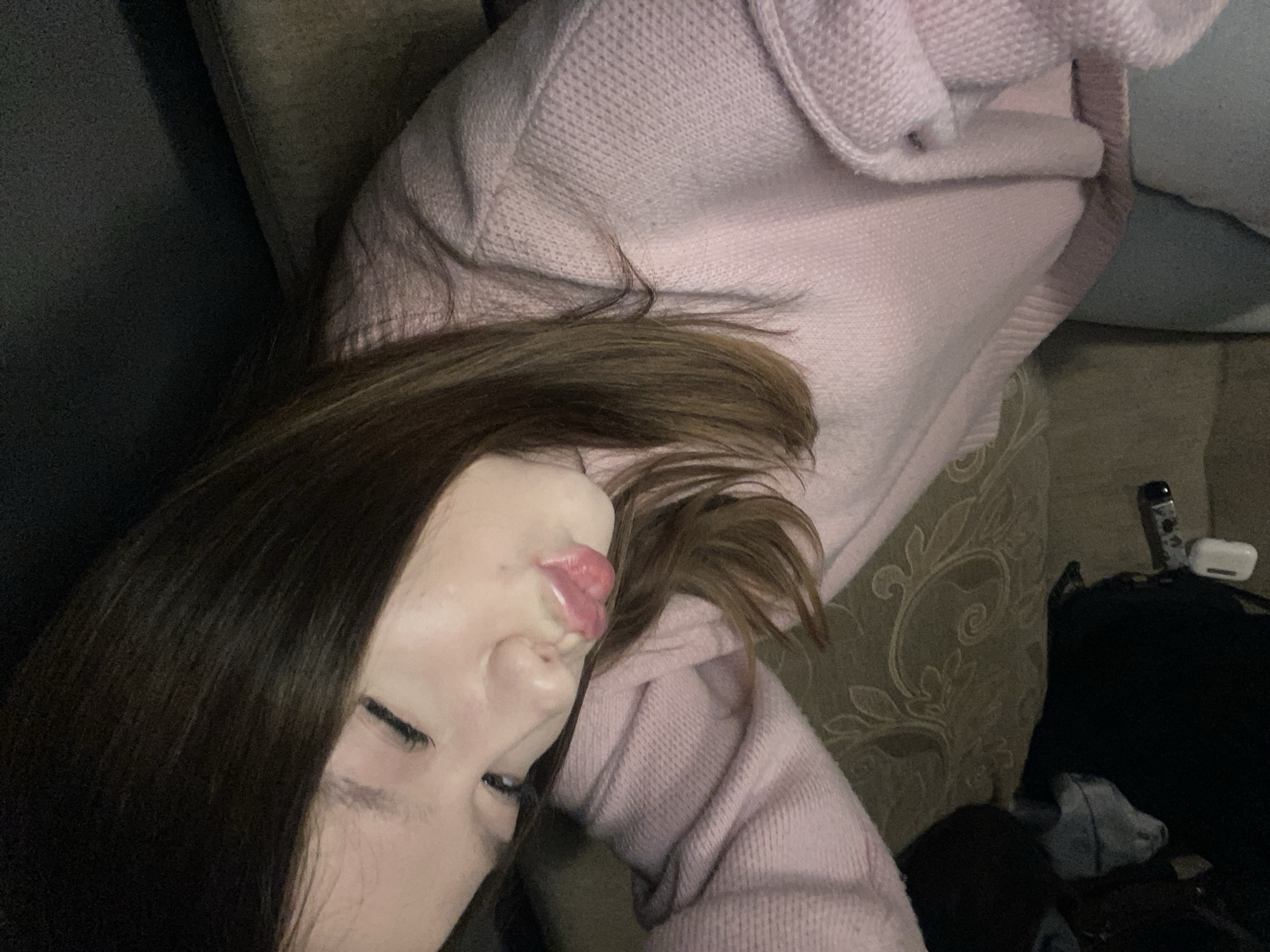 A person with dark brown hair is lying down on a couch. The person is wearing a light pink sweater. The couch has a patterned back. To the right of the person, there is a black bag, a remote control, and a small white object on the couch. The location is unknown.