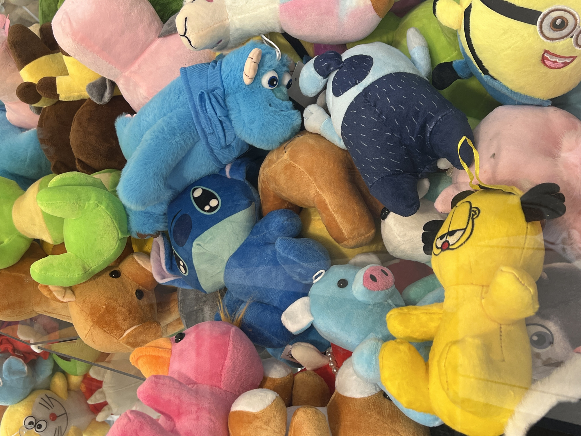 A dense pile of assorted plush toys fills the frame. Prominent figures include blue cartoon creatures resembling "Stitch" and "Sulley" from "Monsters, Inc.", a yellow "Minion", and a yellow Garfield plush. Other plushies include a pink flamingo, brown deer, blue pig, a green limb, and a white lama. The arrangement gives the impression of a claw machine prize pile or a similar display. There is no evidence suggesting the location is in Krasnodar, Russia.
