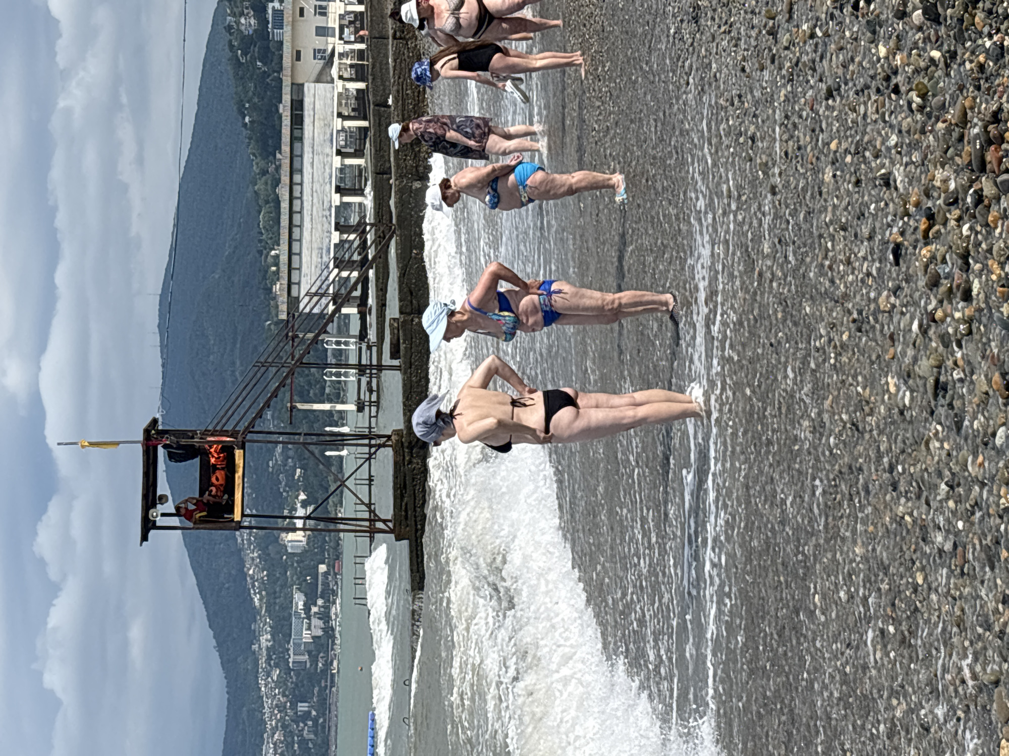 The image depicts a pebble beach in Sochi, Russia, with the Black Sea in the foreground. In the background, there are steep, tree-covered mountains with a city built on their slopes. A lifeguard tower stands on the beach, a structure of metal and wood with a yellow flag at its peak. Several people, mostly women, are visible on the beach, dressed in swimsuits and some wearing headscarves. They appear to be standing in or near the shallow water, with waves breaking on the shore. Some are facing the sea, while others are looking away. A building with a long, low roof supported by pillars is situated on a pier-like structure extending into the sea. The sky is overcast with clouds.