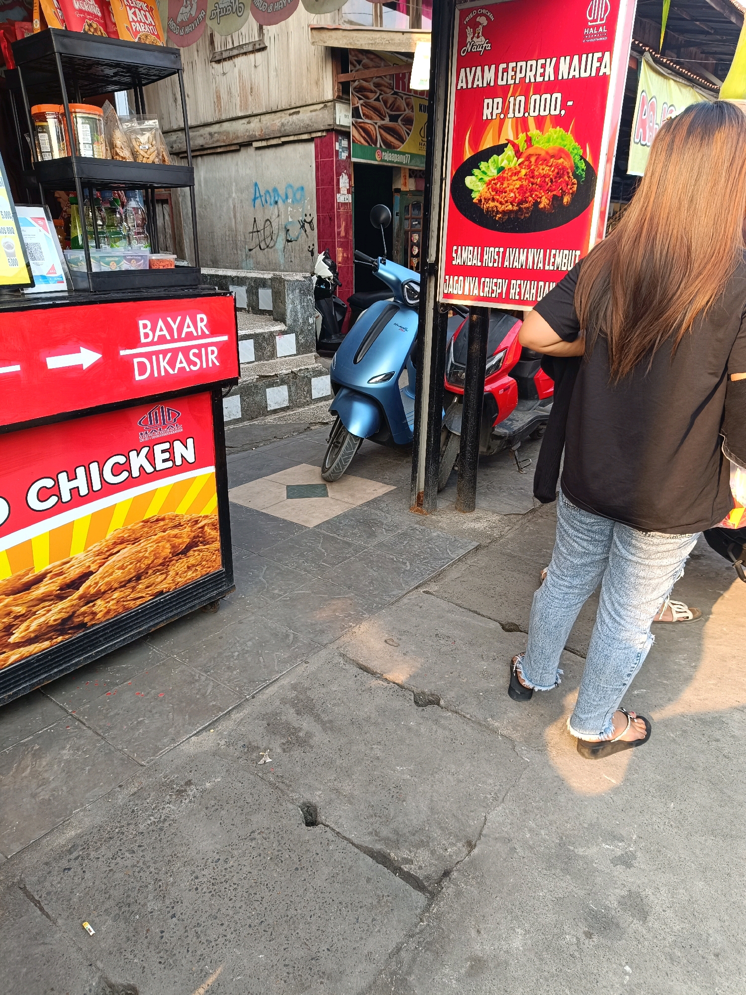 The image depicts a street scene in Kayu Agung, Indonesia, with a focus on a food stall and a woman. The food stall features a prominent red sign advertising "Fried Chicken" and "AYAM GEPREK NAUFA" with a price of "RP. 10.000,-". The stall is stocked with various food items, including packaged snacks and drinks. To the left of the stall, a sign with white text on a red background reads "BAYAR DIKASIR" (Pay at the counter).
A woman with long, light brown hair, wearing a black t-shirt and light-wash, distressed denim jeans, stands facing away from the camera. She is wearing black platform flip-flops. Her posture suggests she is looking at the food stall's offerings or waiting in line.
In the background, two scooters are parked: a bright blue one on the left and a red one on the right, partially obscured by the woman. The environment appears to be an outdoor urban setting with a sidewalk paved with gray tiles and concrete. Graffiti is visible on a wall in the background, and there are some scattered pieces of debris on the ground. The lighting indicates it is daytime, with sunlight casting shadows.