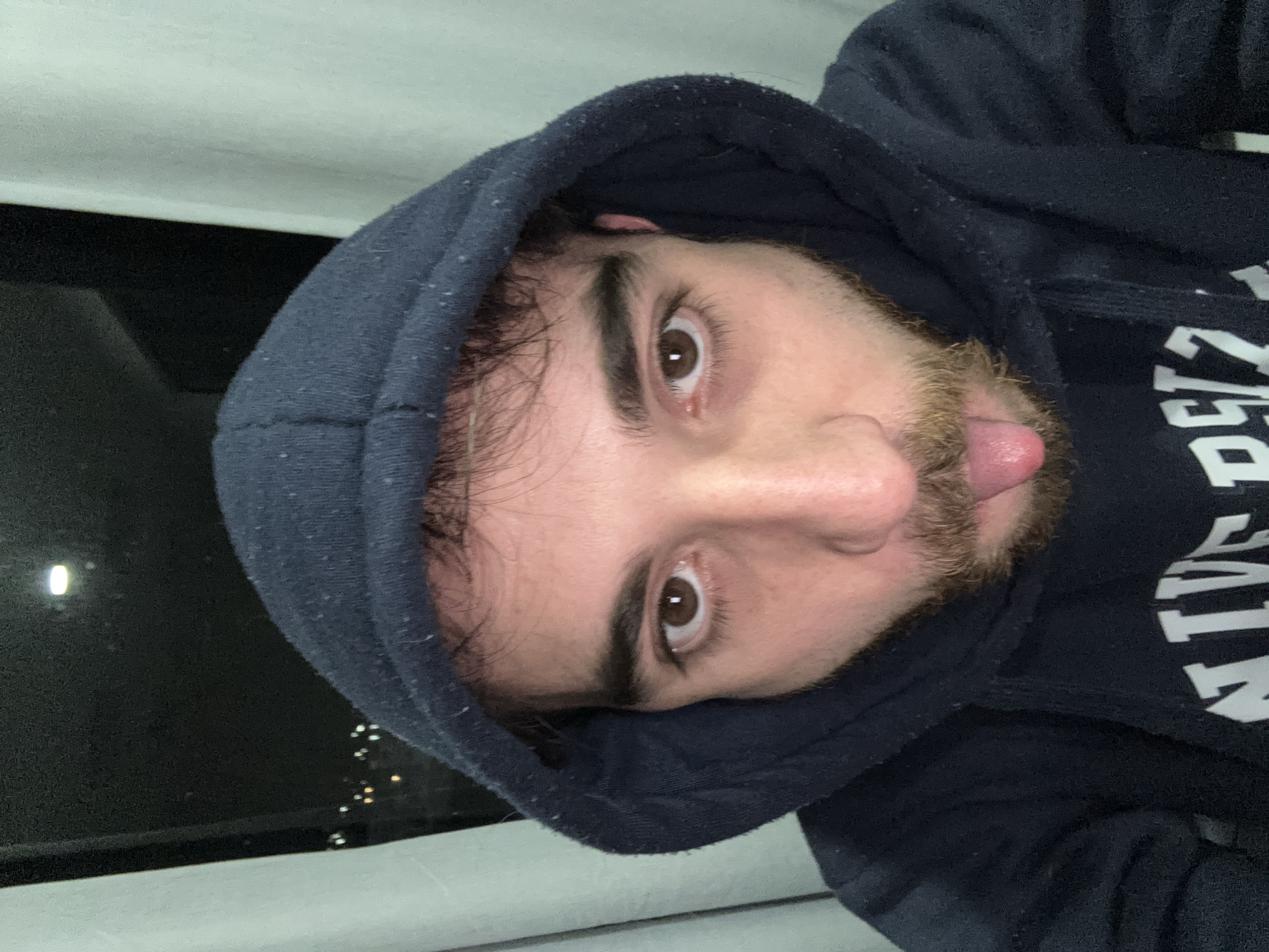 A man with brown eyes and a beard, wearing a dark hooded sweatshirt, is sticking his tongue out. He appears to be inside, looking out a window at night. The window reflects a cityscape, possibly lights from a distant area. The location is unknown.