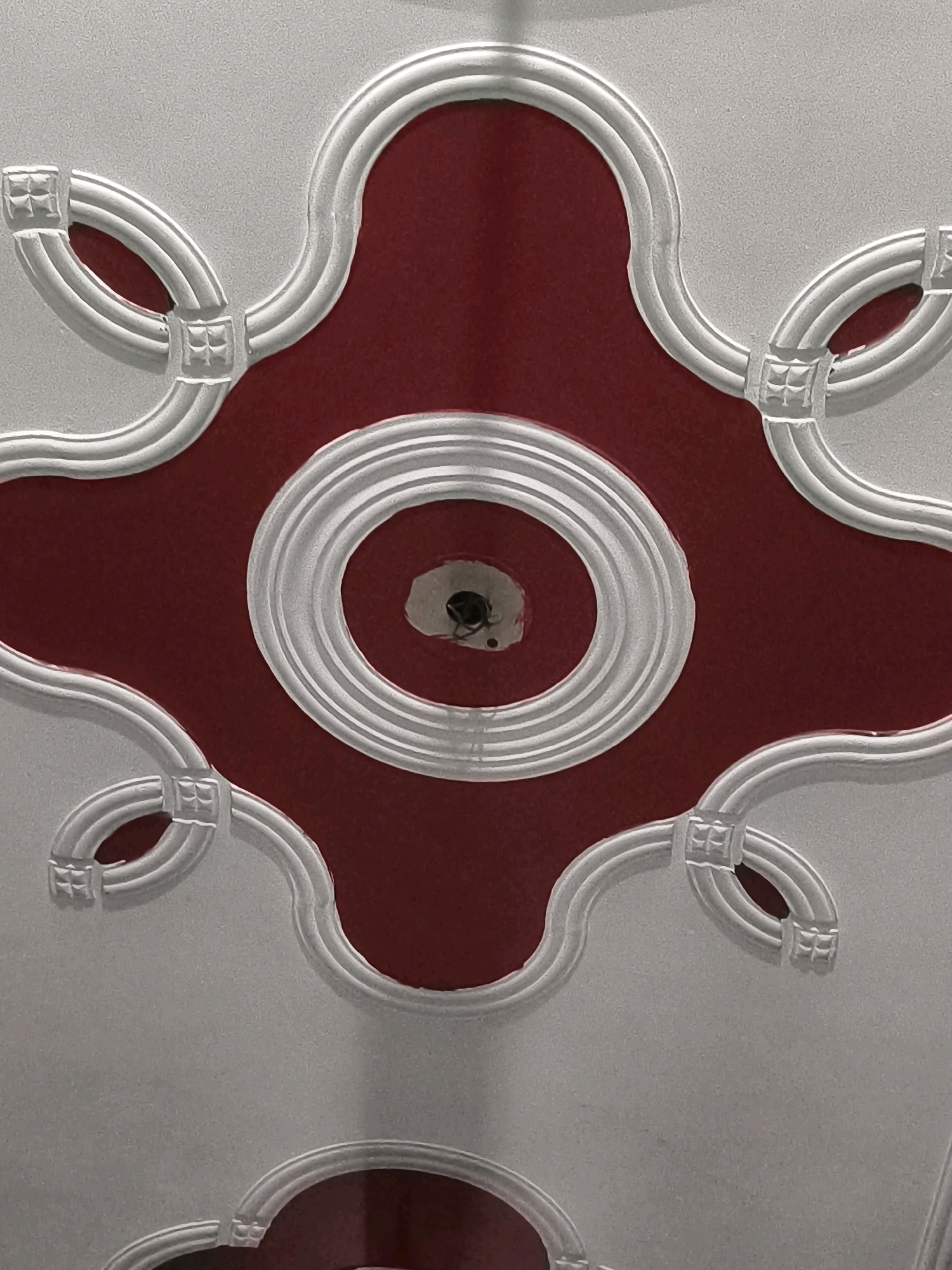 The image shows a close-up view of a decorative ceiling with a central element featuring concentric rings and a hole in the middle. The ceiling is painted in a two-tone design, with a deep red color filling the central area and a silver-grey color on the surrounding decorative elements. These elements are elaborate, featuring raised white molding with a symmetrical pattern of curved lines and square-shaped embellishments. The overall design suggests an ornate interior, likely in a residential or commercial building. There are no people visible in the image. The specific location in Ghaziabad, India, is not discernible from the visual information alone.