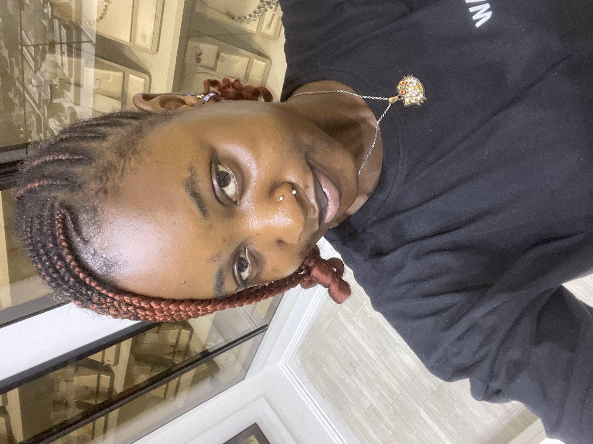 The image shows a close-up, rotated selfie of a person in Unguwar Sarki, Nigeria. The subject has dark skin and braided hair with reddish-brown highlights. They are wearing a black long-sleeved shirt and a silver necklace with a small, colorful pendant that resembles a Hello Kitty character. The person also has a nose ring and an earring. Their expression appears neutral.
The background suggests an indoor setting, possibly a store or a home, with display shelves visible behind the subject, filled with what appear to be jewelry or small decorative items. The lighting is soft and likely artificial, indicating it could be daytime or evening. The overall ambiance is casual and personal. There is some text visible on the shirt, partially obscured, which appears to be "W M".