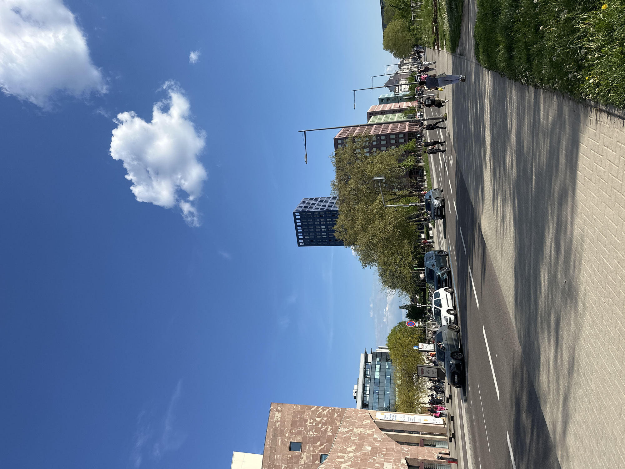 The image shows a street scene in Strasbourg, France, characterized by modern architecture, vehicular traffic, and pedestrian activity under a bright blue sky with a prominent white cloud.
Buildings: A mix of architectural styles is visible, including brown and tan stone-clad buildings. There are also modern buildings with green and red window panels and a taller, dark-colored skyscraper with a grid-like facade.
Transportation: Cars are parked and moving along the street, with white lane markers visible. Pedestrians are walking along the sidewalk.
Environment: The scene is well-lit with clear weather conditions. There are trees lining the street, and a patch of grass visible along the sidewalk. Shadows cast by trees stretch across the pedestrian area.
People: Pedestrians are walking on the sidewalk; their actions seem to be casual and everyday.
Objects: Streetlights, road signs, and advertisements, plus vegetation such as trees and patches of grass.
Actions: Vehicles are driving, and people are walking.
Interactions: It depicts people moving along the road.