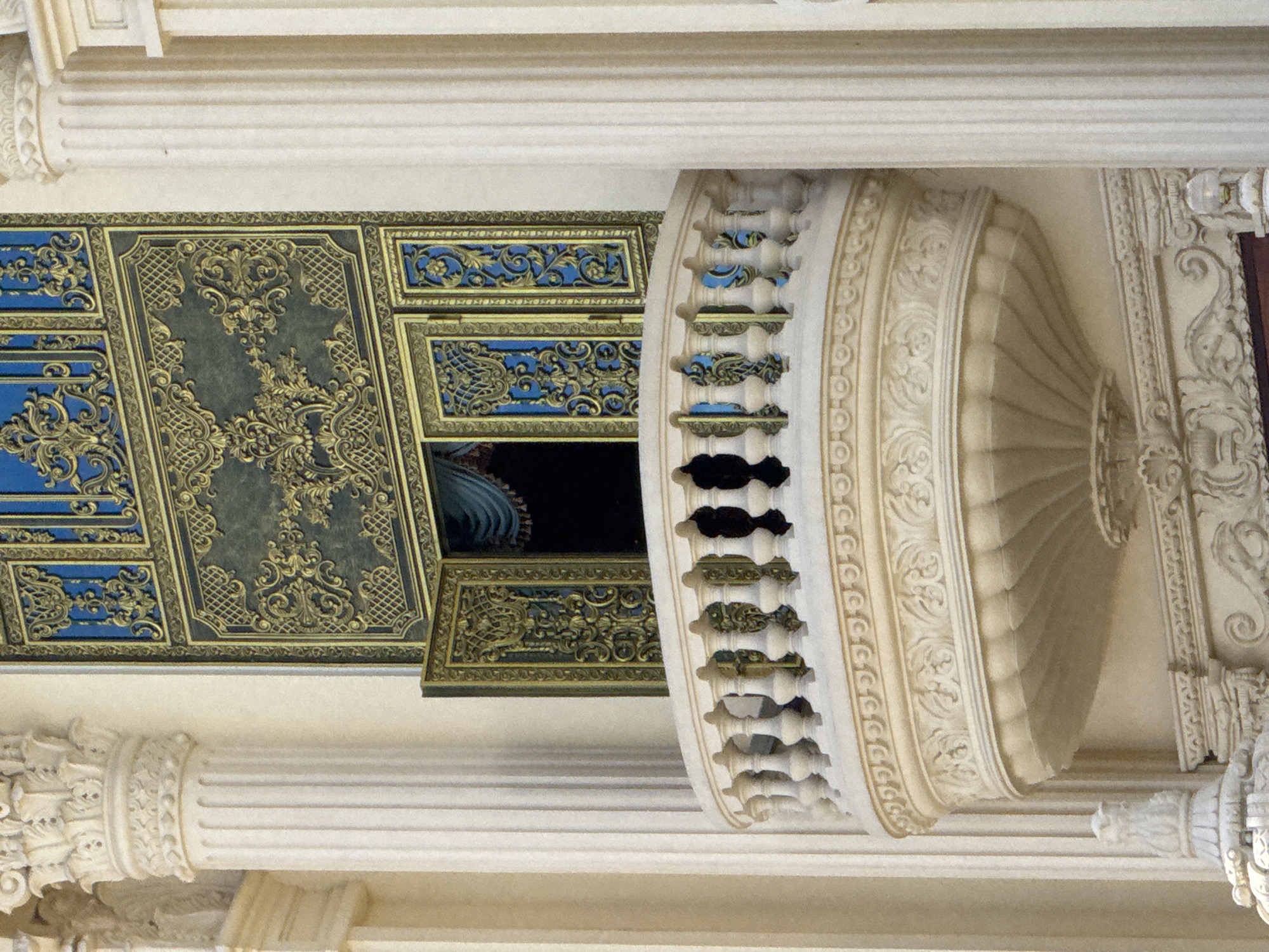 The image captures a portion of a building's facade, featuring ornate architectural elements. A cream-colored, semicircular balcony with a decorative railing and intricate stonework extends from the building. Behind the balcony, a large, elaborately decorated window or door is visible, with sections of deep blue and gold ornamentation featuring floral and geometric patterns. One side of the window is open, revealing a glimpse of blue drapery. Two large, fluted cream-colored columns frame the scene on either side. The building's facade appears to be made of a light-colored material, possibly stone or stucco. The overall style suggests a grand, classical, or baroque influence. The image is focused on the architectural details and does not include any people or specific environmental context beyond the building itself. This ornate balcony and facade are part of the entrance to the Thien An Monastery in Da Lat, Vietnam.
