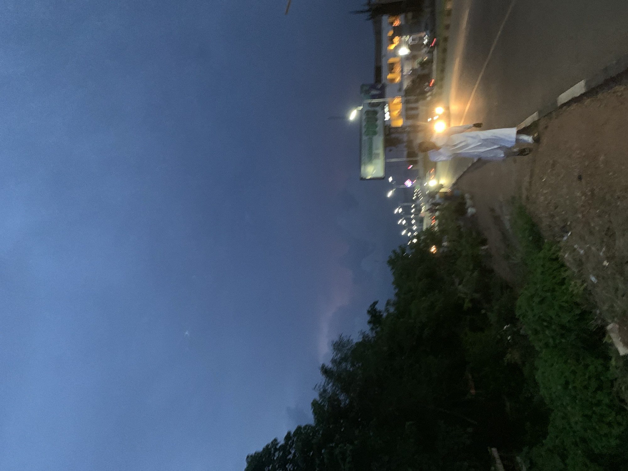 The image captures a scene on a road in Jos, Nigeria, during twilight. A lone individual, dressed in a white robe and headscarf, walks along the shoulder of a paved road. The road is illuminated by streetlights and the headlights of passing vehicles. In the background, buildings with lit windows and a large billboard with green text are visible. To the left of the road, dense green foliage forms a natural barrier. The sky is a deep blue, indicative of the fading light.