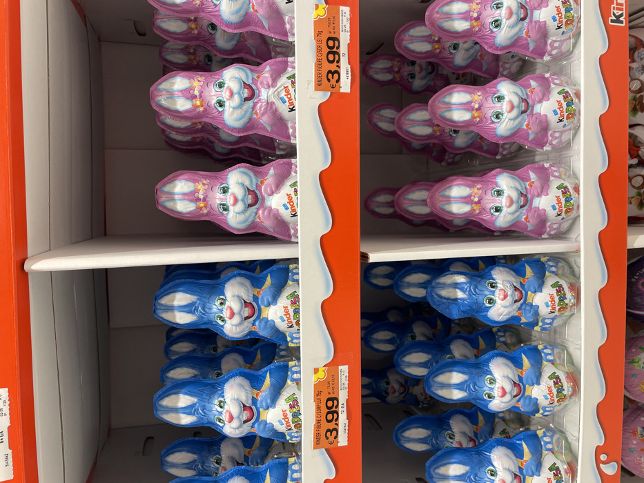 A retail display within a supermarket in Monsummano Terme, Italy, exhibits multiple rows of foil-wrapped chocolate bunnies. The subjects are Kinder Sorpresa brand chocolate figures, specifically "Kinder Figure C/Sorp Lui 75g" as indicated on the price labels.
The display unit features white cardboard dividers and a prominent red header shelf trim. Two primary types of chocolate bunnies are presented:
1. **Blue Bunnies:** These figures are predominantly blue and white, depicting male rabbits with cheerful expressions, holding a Kinder Surprise egg. They are arranged in rows on white display trays within the shelving unit.
2. **Pink Bunnies:** These figures are predominantly pink and white, depicting female rabbits adorned with floral headbands, also with cheerful expressions and holding a Kinder Surprise egg. They are similarly arranged in rows on white display trays, separated from the blue bunnies by a vertical divider.
Each type of bunny is stacked in at least two visible tiers. Price tags are affixed to the red trim of the shelf, clearly displaying "€3.99" for the 75-gram figures. Additional textual information on the tags includes product codes and further pricing details. The environment is illuminated uniformly, consistent with indoor commercial lighting. No individuals or active interactions are present in the frame; the scene solely depicts a static product display.