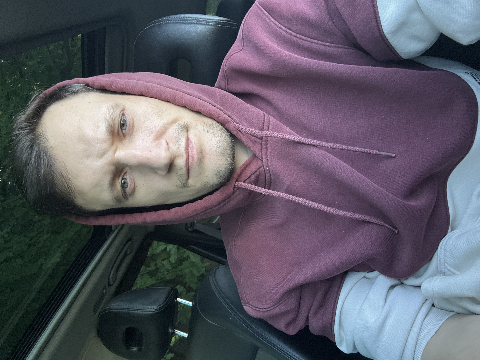 A man with fair skin, light brown hair, and light green eyes is sitting in the driver's seat of a car, facing the camera. He is wearing a maroon hoodie with white sleeves and a hood pulled up around his head. His expression is neutral. The interior of the car is visible, with black leather seats and a sunroof. The background appears to be trees and greenery, suggesting an outdoor location, possibly a rural area or a park. The overall lighting suggests it is daytime, but the exact time is unclear. No specific location within Makiv, Ukraine, can be identified from the visual information provided.