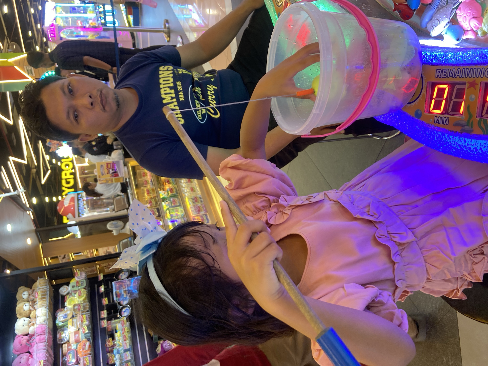 In Sidoarjo, Indonesia, a young girl in a pink ruffled dress and a blue polka-dot headband is playing a fishing game at an arcade. She holds a fishing rod with a string and a yellow float, attempting to catch something in a clear plastic bucket filled with water. Next to her, a man wearing a dark blue t-shirt with "CHAMPIONS NBA 2015 GOLDEN STATE CURRY" printed on it, sits with his arms resting on his lap, observing. The background reveals an arcade environment with shelves stocked with stuffed toys and various prizes, along with illuminated game machines, characteristic of entertainment venues in Sidoarjo. The lighting in the scene is bright and colorful, with prominent purple and blue neon lights from the game machine.