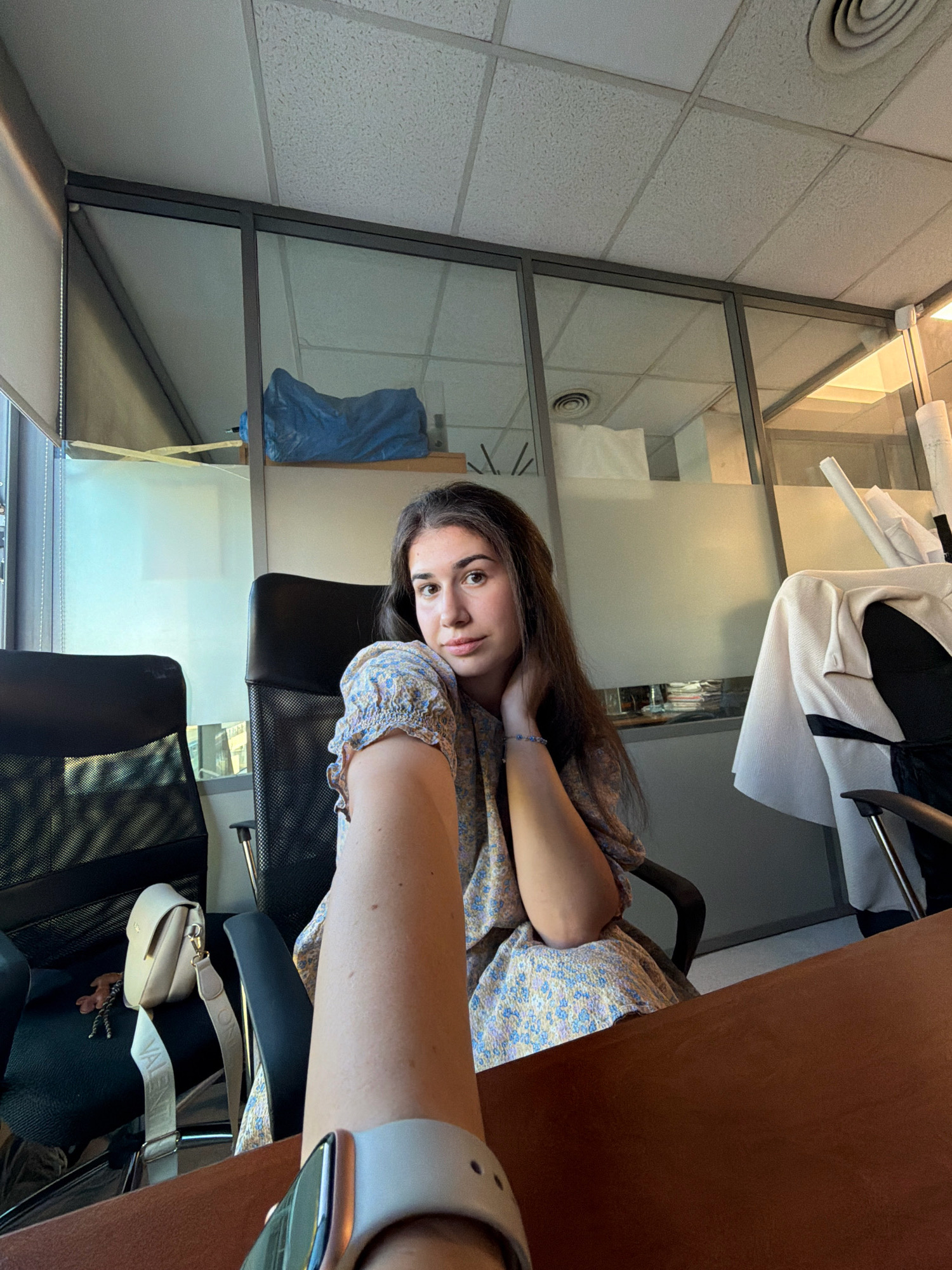The image shows a young woman sitting in an office. She is wearing a light-colored floral dress and has long, dark hair. She is looking at the camera and resting her head on her hand. In the background, there are glass walls, office chairs, and a desk. The scene appears to be an office in Sofia, Bulgaria, suggested by the presence of a specific type of ceiling tile and air vent common in buildings in Sofia, and the overall lighting and window style typical of an office building in the city. A purse with a distinctive strap is visible on the chair to her left. A person's arm wearing a smartwatch is in the foreground, indicating a selfie-style shot.