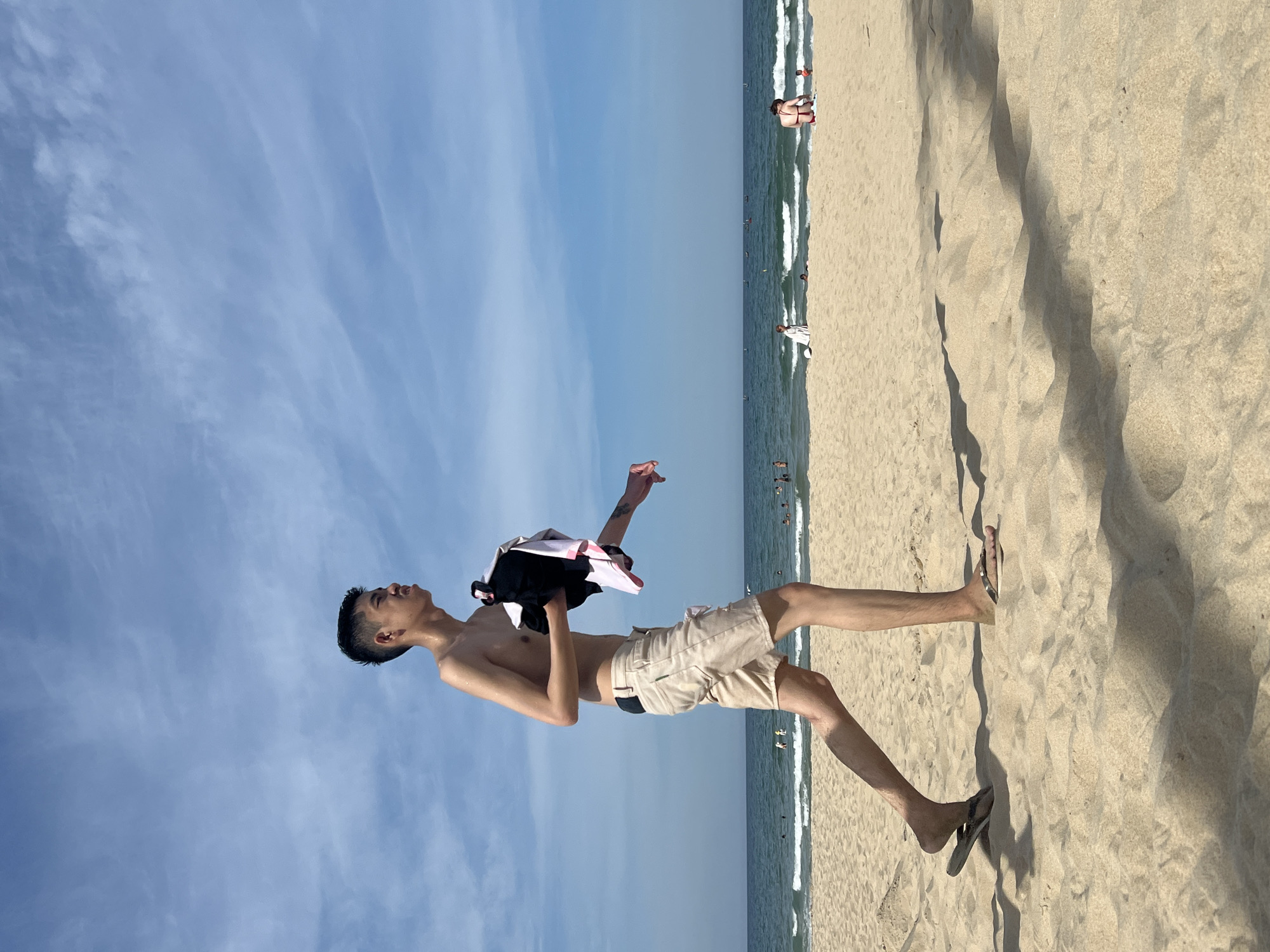 In Đà Nẵng, Vietnam, a young, shirtless man with dark hair walks across a sandy beach. He is wearing light-colored shorts and flip-flops and carries a black and white garment in his right hand. The man's body is angled to the left, and he appears to be looking towards the sea, which is visible in the background with gentle waves. The sky is blue with scattered white clouds. Several other beachgoers are present in the distance, some in the water and others on the sand, indicative of a public recreational area. The sand is light brown and shows footprints and shadows from nearby objects, suggesting a sunny day.