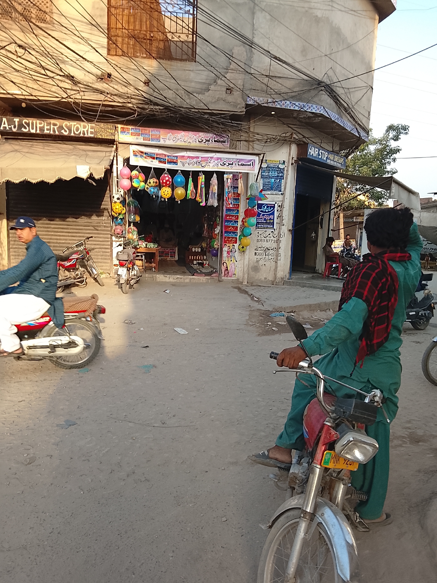 A street scene in an urban area in Unknown, Unknown is captured in this outdoor photograph. The image includes individuals, vehicles, and commercial establishments under an overcast sky.
In the foreground, a person dressed in green sits on a red motorcycle with a silver front wheel, facing away from the camera. The motorcycle's license plate is visible. To the left, another person in a blue shirt and white pants rides a similar red motorcycle in the same direction.
The background features a row of shops with signage in a non-English script. The leftmost shop is named "A-J SUPER STORE", and the adjacent shop has balloons and toys displayed outside. Further down the street is another establishment named "HAIR STOP HAIR DRESSERS".
Several motorcycles are parked in front of the shops, and people are seen inside and near the shops. Overhead, a dense network of electrical wires creates a chaotic pattern against the buildings. The street appears to be paved, with some patches of discoloration. The lighting suggests it may be late afternoon or early morning.