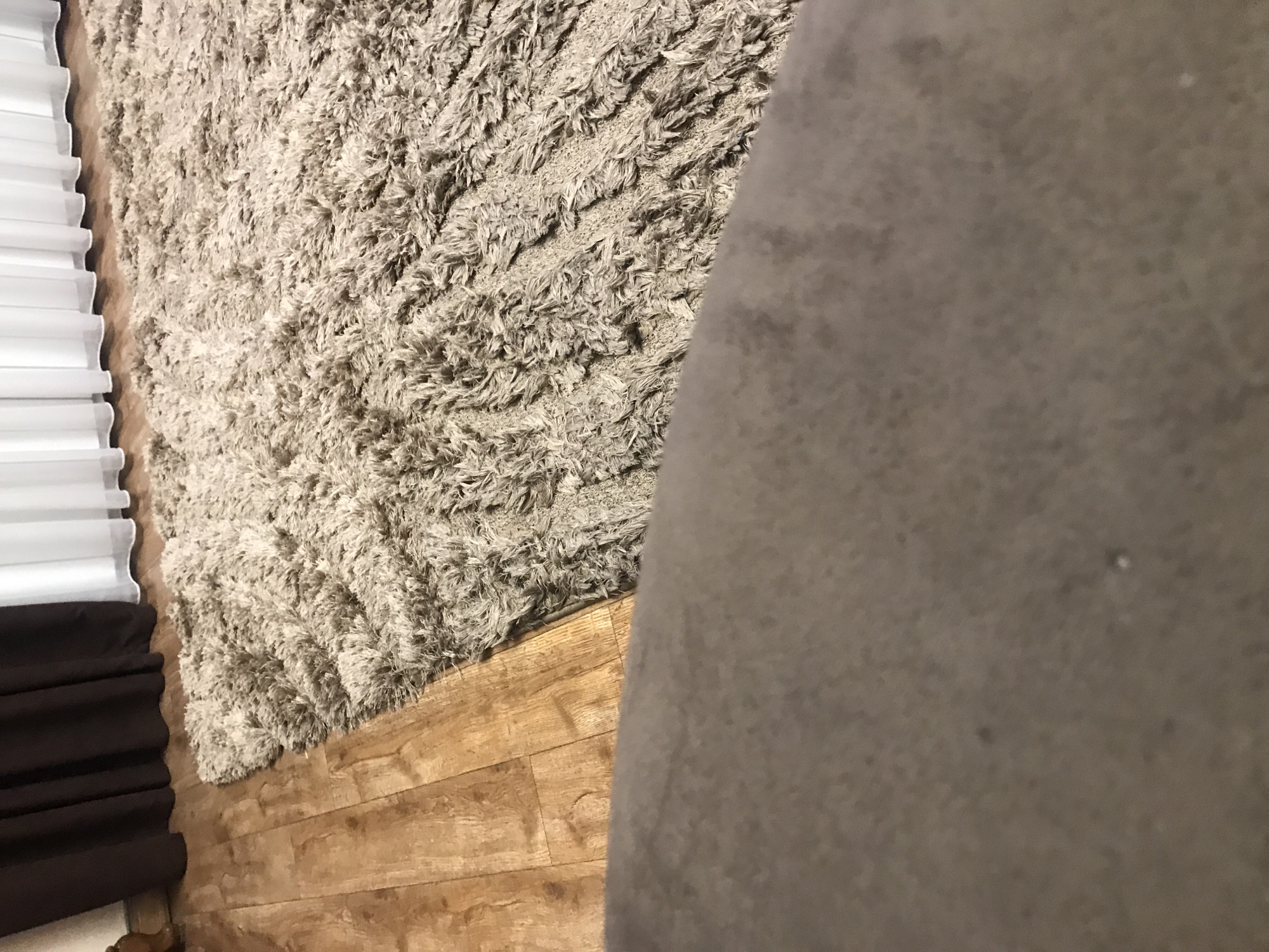 The scene is a close-up, low-angle shot inside an unidentified building. The flooring is composed of tan-colored, wood-grain planks. Partially visible on the left are white and brown pleated blinds. Adjacent to the blinds, a beige shag rug occupies a significant portion of the frame. A large, gray, textured object dominates the right side of the image. The location in Yerevan, Armenia, cannot be determined from this information.