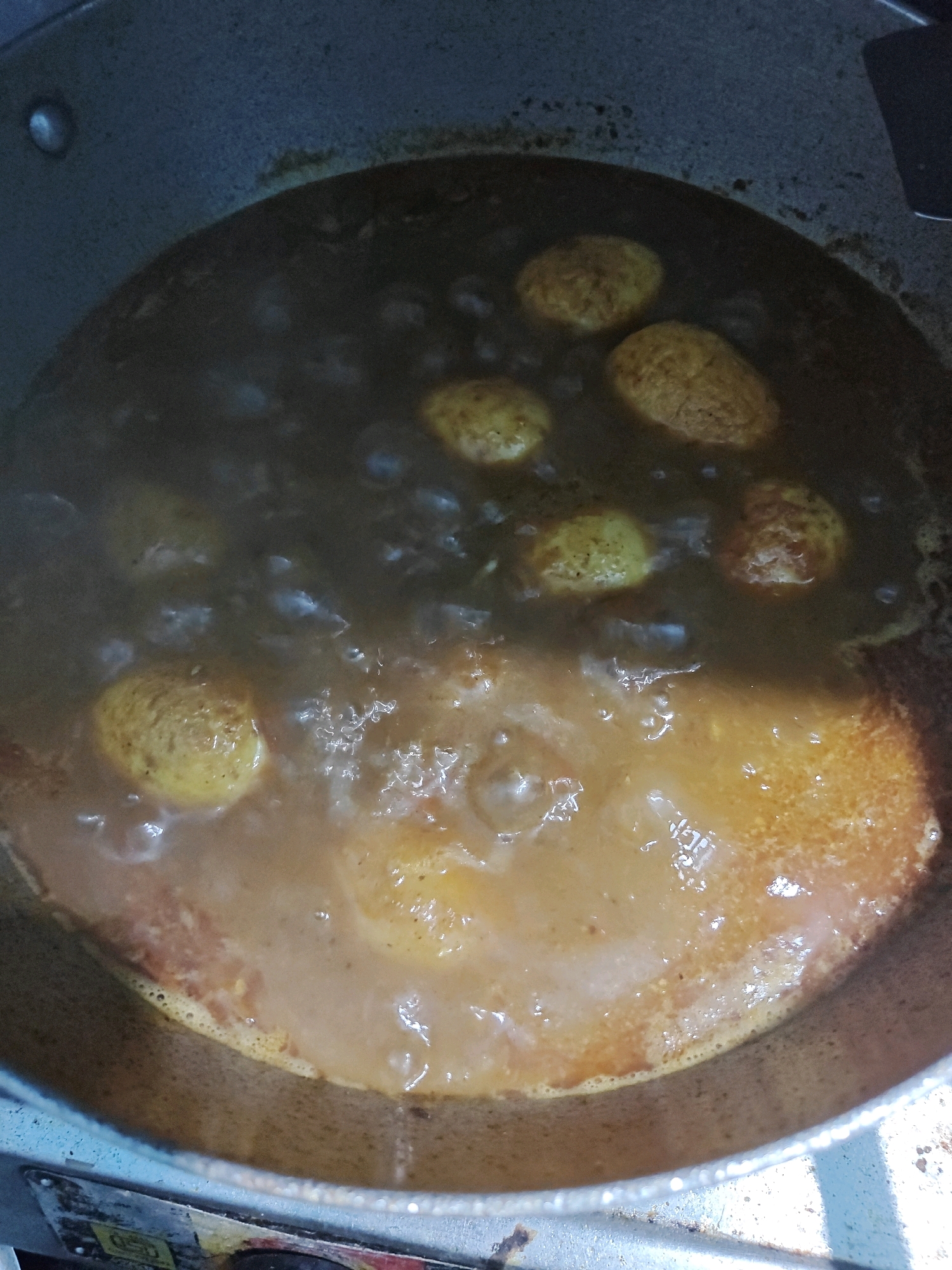 The image shows a close-up view of a pot of food being cooked, likely a curry with boiled eggs. The food is simmering in a rich, brown sauce with visible bubbles. Several whole boiled eggs, with a yellowish hue, are submerged in the sauce. The pot itself appears to be made of metal and has a slightly worn appearance. The lighting suggests an indoor setting, possibly a kitchen. There are no people visible in the image. The lower part of the image shows a portion of what appears to be a stove or cooking surface, with some wear and tear. There is no visible indication of the specific location within Ranchi, India, beyond it being a cooking scene in a domestic or commercial kitchen.