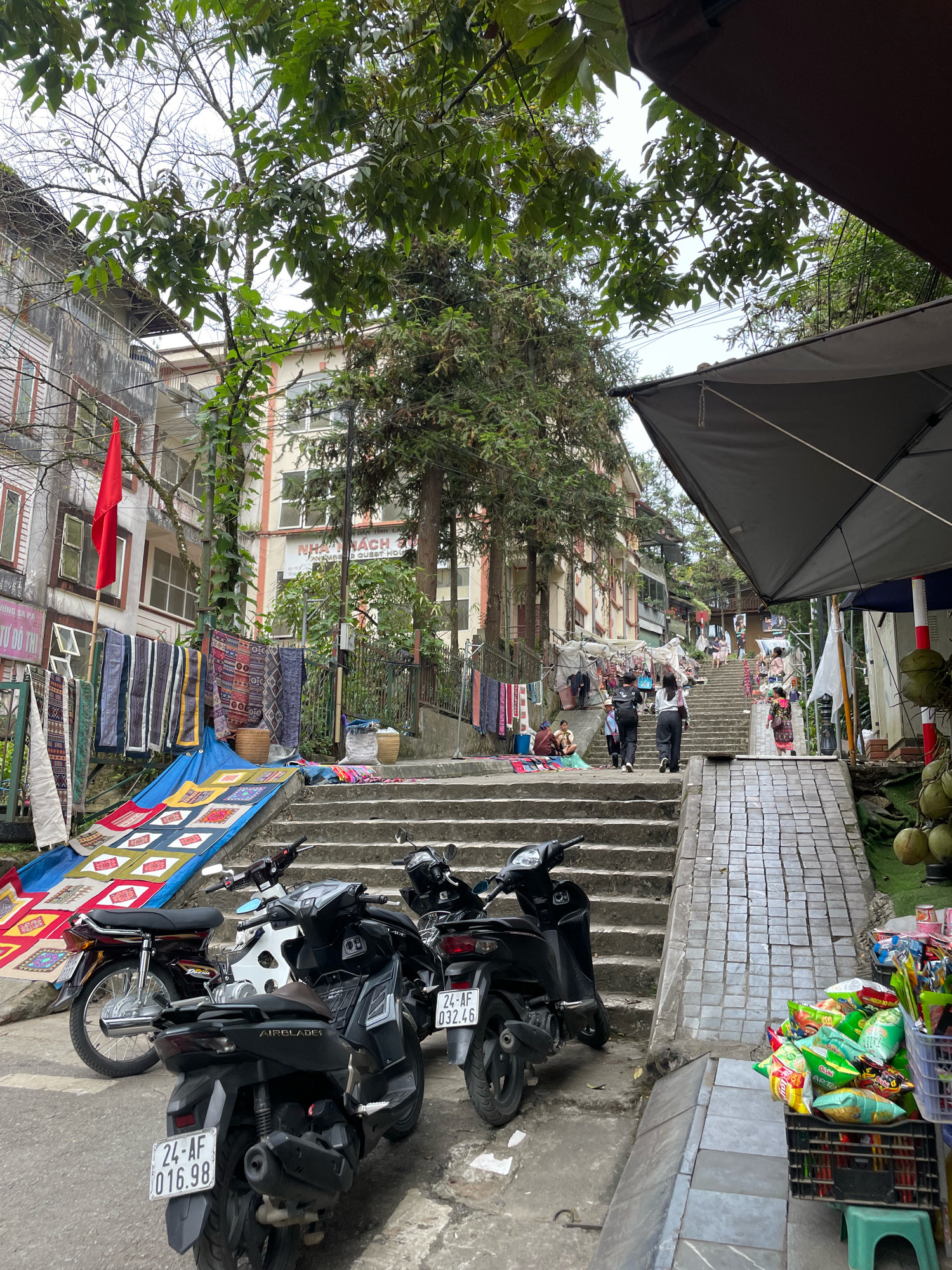 In Sa Pa Ward, Vietnam, a street scene unfolds with a focus on a steep, outdoor staircase. The architecture suggests a bustling commercial area, with buildings lining the sides of the ascent. On the left, a vibrant display of textiles, possibly for sale, is laid out on a blue tarp, featuring colorful patterns and intricate designs. Several motorcycles are parked on the street, their presence indicating the common mode of transport in the area.
As the eye moves up the staircase, several individuals can be observed ascending and descending. Some are tourists, identifiable by their casual attire and backpacks, while others appear to be locals, engaged in daily activities. The atmosphere seems lively, with vendors possibly operating stalls along the route, as suggested by the baskets and merchandise visible. The overall impression is one of a dynamic, outdoor marketplace set against the backdrop of a hilly urban landscape. The foliage from trees interspersed throughout the scene provides a natural element to the otherwise man-made environment.