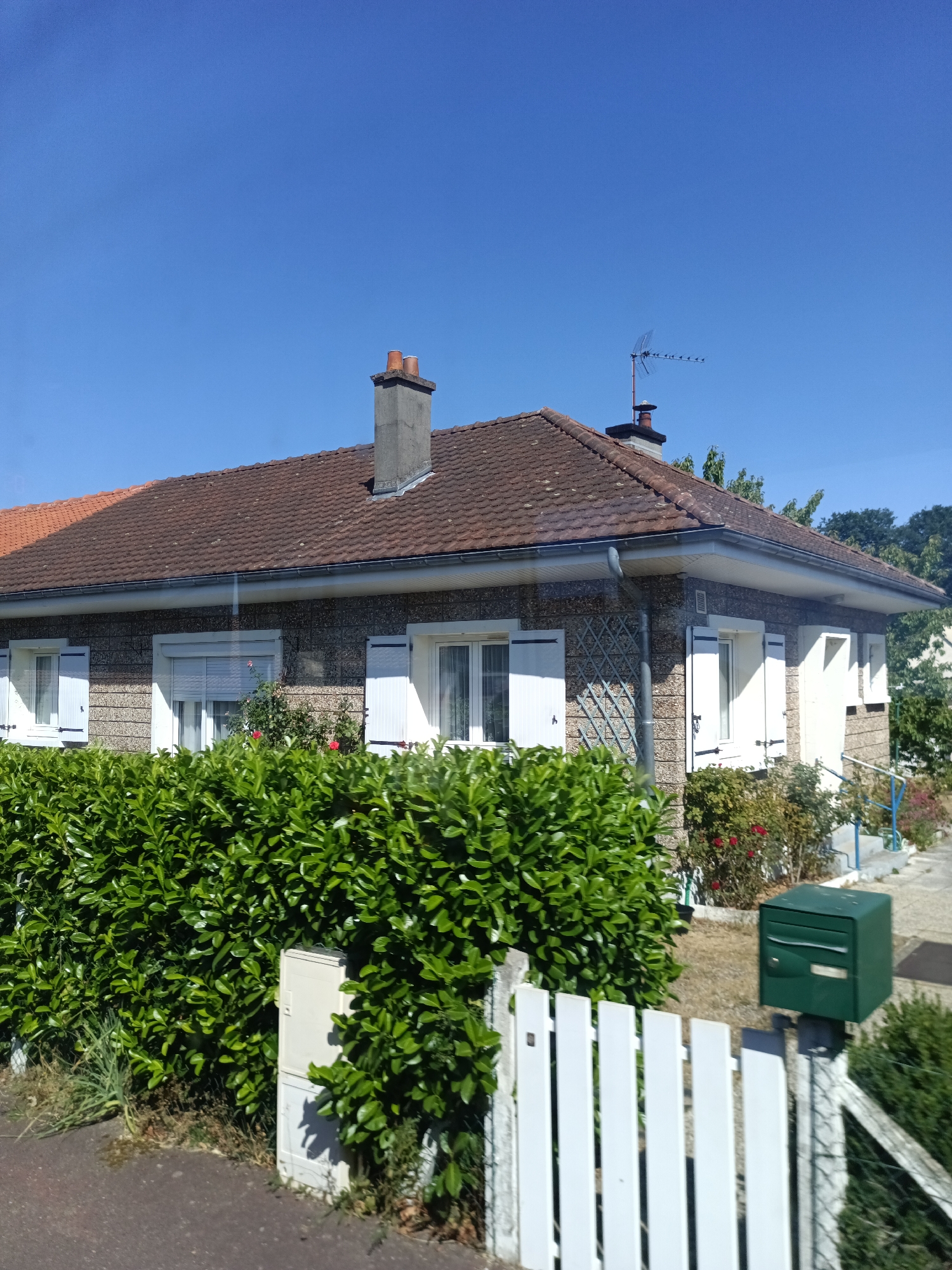 In Yainville, France, on a clear day, a low-rise house with a brown tiled roof is visible. The house has a speckled facade and features white-shuttered windows and a white door. A dense green hedge runs along the front of the property, obscuring the lower part of the house. Beyond the hedge, a white picket fence borders the scene, and a green mailbox stands to the right. A chimney with two visible flues rises from the roof. An antenna is also visible on the roofline. The background reveals trees and a bright blue sky.