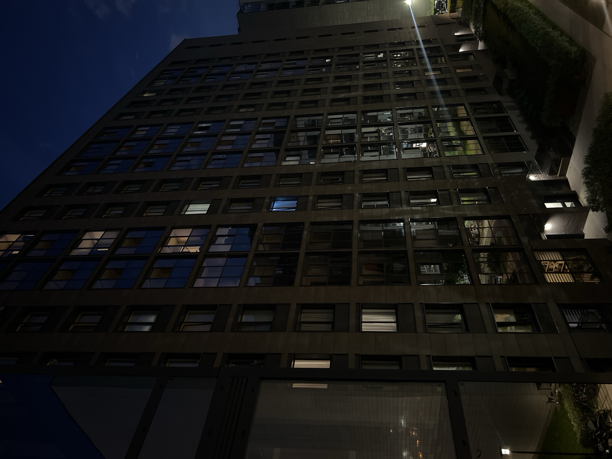 The image captures an exterior view of a modern, multi-story apartment building at dusk or night. The building is constructed with a combination of light-colored facade panels and dark, possibly glass, balconies or window frames. Many windows are illuminated from within, suggesting residents are present. The building is situated in Sinyavino, Russia. In the foreground, there is a sidewalk or pathway adjacent to a well-maintained green lawn and dense shrubbery. A streetlamp illuminates a portion of the pathway. Bicycles are parked on the sidewalk. In the background, other apartment buildings and a dark sky with visible clouds are present. The overall impression is one of an urban residential setting at the end of the day.