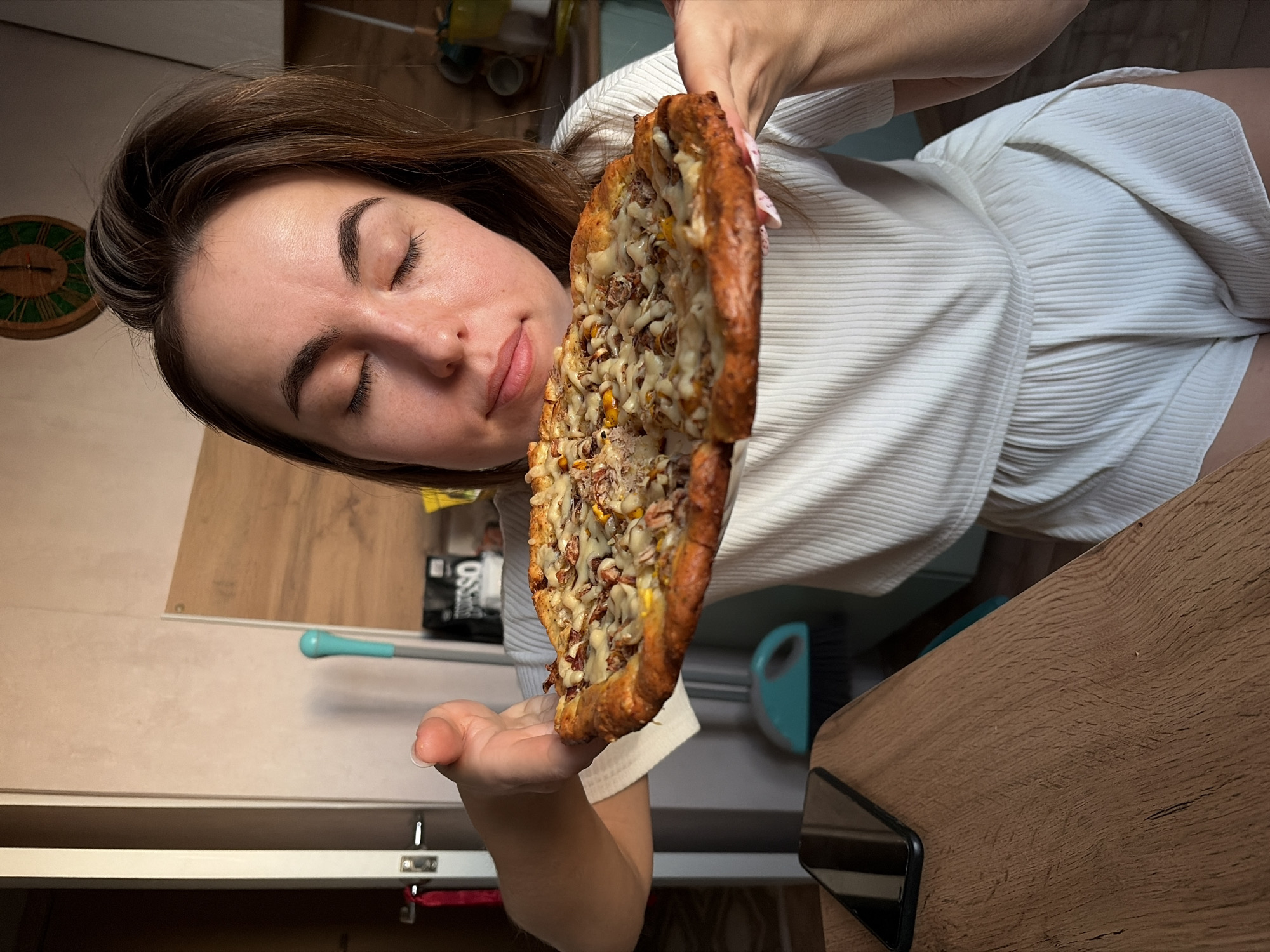 A woman is holding a pizza up to her face. She is indoors, in a kitchen. The woman has brown hair and is wearing a white shirt and shorts. She has her eyes closed and her mouth slightly open as if she is about to eat the pizza. The pizza is round and has a brown crust with cheese, meat, and corn on top. The kitchen has light-colored cabinets and a wooden countertop. There is a clock hanging on the wall, as well as a broom. The location is inferred as Izhevsk, Russia.