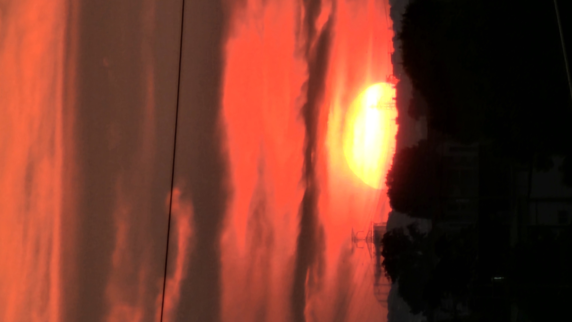 This is a vertical, full shot of a sunset over Asan-si, South Korea. The sun is a bright, yellow orb with a soft halo, partially obscured by dark, silhouetted clouds that stretch across the frame. The sky is a vibrant gradient of red and orange, with some darker, more ominous clouds interspersed. In the foreground, a dark silhouette of trees, buildings, and a power line are visible against the dramatic sky. The overall impression is one of dramatic natural beauty, with a touch of urban interference.