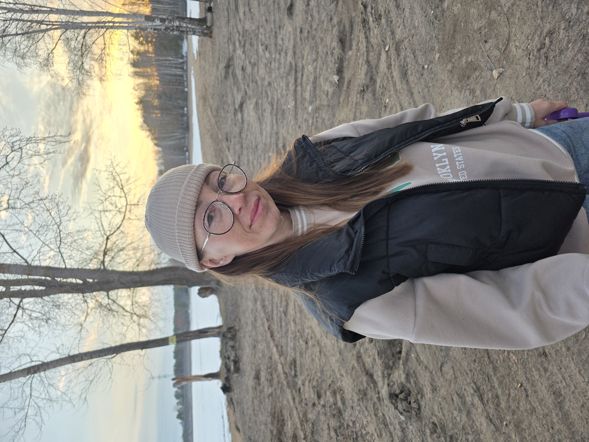 A woman with fair skin, wearing glasses and a beige beanie, is partially depicted in a landscape featuring sand and trees, likely taken in Noviy Urengoy, Russia. The woman is wearing a beige sweatshirt with the word "BROOKLYN" and "UNITED STATES" printed on it, layered under a black vest. She is holding a purple object in her hand. The background shows bare trees and a body of water with reflections of the sky, suggesting it might be a lake or a river during the late afternoon or early evening, judging by the warm light of the sunset. The ground appears to be sandy with some vegetation. The orientation of the photo is rotated counterclockwise.
