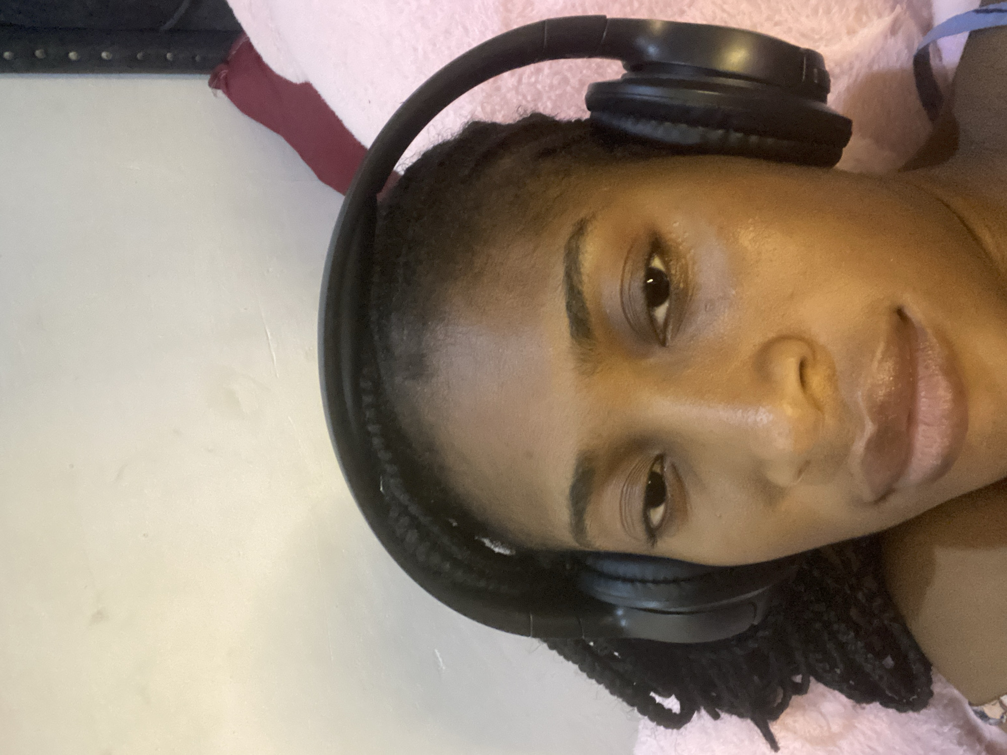 The image shows a close-up, frontal, selfie-style shot of a Black woman wearing black over-ear headphones. She has braided hair. She appears to be lying down, as evidenced by the soft, pink fabric in the background, which could be a blanket or pillow. A dark red piece of fabric is visible in the upper right quadrant of the image. The background wall is light-colored and textured. The lighting is warm, suggesting an indoor setting. The woman's expression is neutral, and she is looking directly at the camera. The image was captured in Kubwa, Nigeria.
