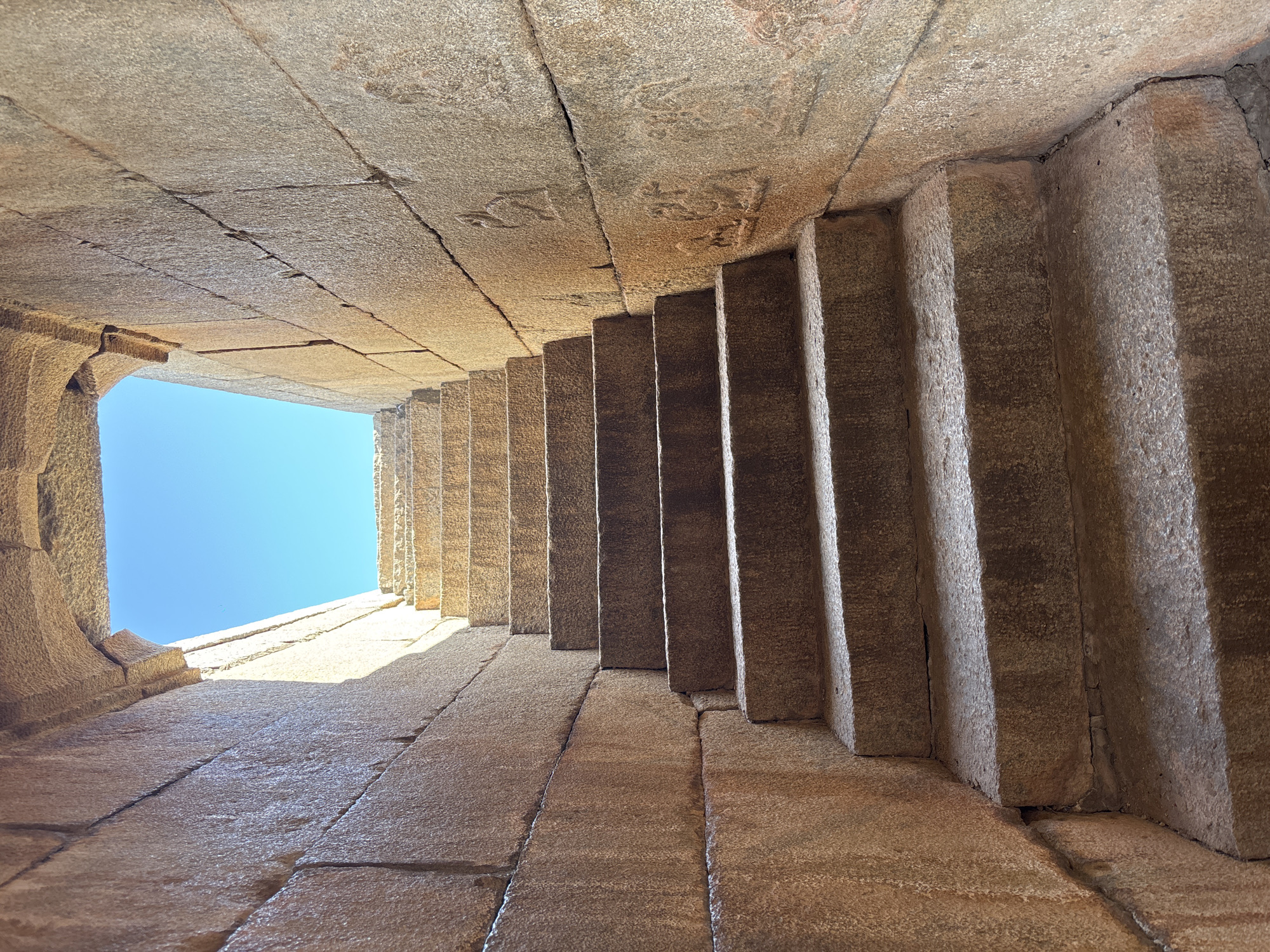 The image depicts a set of stone steps leading upwards towards an opening with a view of the blue sky. The structure is made of large, roughly hewn stone blocks, with cracks and irregularities visible. The steps are vertical and narrow. The opening at the top is square and appears to be the only source of natural light. This is likely a structure found in Kamalapura, India, which is known for its historical stone architecture. There are no people visible in the image. The environment appears to be dry and arid.