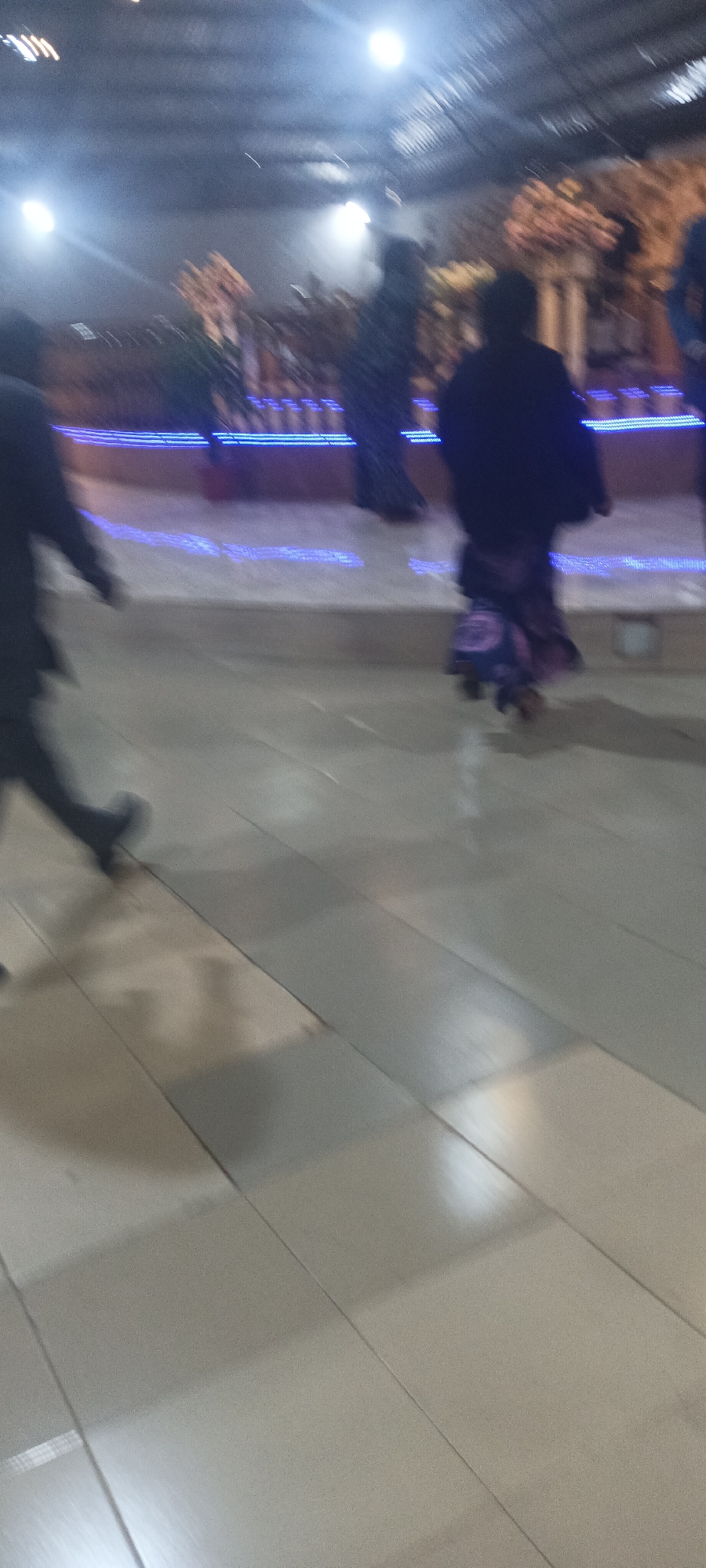 The image depicts a low-light indoor or semi-outdoor venue on Rukuba Road, Nigeria, characterized by significant motion blur. Multiple individuals are in motion across a light-colored, reflective tiled floor. On the far left, the lower body of a person, dressed in dark clothing and shoes, is captured mid-stride, moving towards the right. In the center-right, another individual is observed from behind, wearing a dark upper garment and a patterned skirt or dress with purple and lighter hues, also exhibiting motion towards the right. Further indistinct, shadowy figures are present in the blurry background. A raised platform or barrier extends horizontally across the mid-ground, featuring multiple parallel lines of bright blue LED-like lighting along its edge. Beyond this illuminated barrier, decorative elements, possibly artificial plants or architectural features, are vaguely discernible. Overhead, several bright, circular artificial light sources are visible, casting intense illumination, generating lens flares, and reflecting off a textured ceiling, which appears to have a corrugated surface.