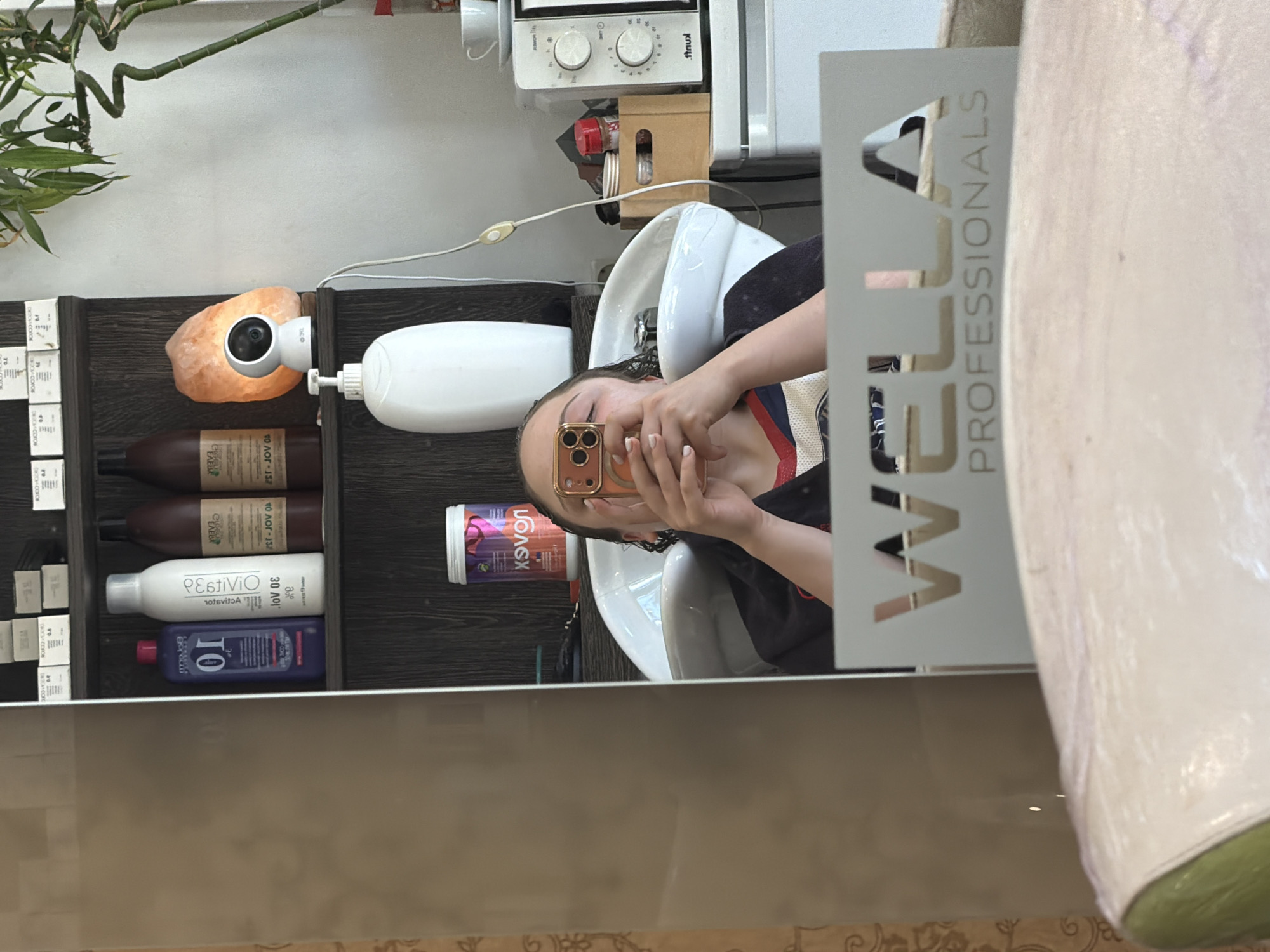 Here is a detailed description of the image:
**General Scene:**
The image appears to be a selfie taken in a hair salon, possibly in Arroyo de la Miel-Benalmádena Costa, Spain. The reflection in the mirror shows a person sitting in a hair salon chair, getting their hair washed in a salon sink.
**People:**
* There is a person visible in the mirror's reflection holding up a cell phone and likely taking the picture. Only the person's head and hands are fully visible.
**Objects:**
* **Hair Salon Sink:** A white, ceramic hair salon sink is present, where the person is having their hair washed.
* **Hair Products:** Shelves with various hair products, including shampoo and styling products, are visible behind the sink. Some product names/brands can be partially read, such as "Kolex" and "PEstiVio."
* **Mirror:** The scene is viewed through a large mirror with a logo.
* **Logo:** A logo for "Wella Professionals" is partially visible on the mirror's right side.
* **Cell Phone:** The person is holding a gold-colored cell phone (likely an iPhone) to take the selfie.
* **Light Fixture:** An orange salt rock with a white camera light is placed on the top shelf.
**Environment:**
* **Hair Salon:** The environment is a typical hair salon setting, with hair washing equipment and product displays. The location of the salon may be in Arroyo de la Miel-Benalmádena Costa, Spain.
**Action:**
* A person is getting their hair washed in the salon.
* The person is taking a selfie in the mirror.