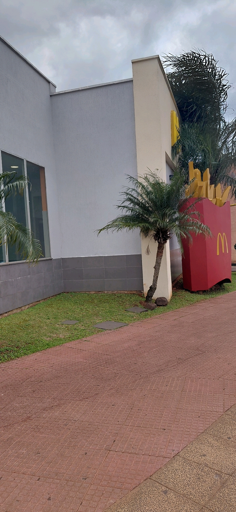 The image depicts the exterior of a McDonald's restaurant in Encarnacion, Paraguay. The building has a light-colored facade with a gray tiled lower section. A large, red McDonald's french fry container sculpture, complete with yellow french fries and a yellow McDonald's logo, is prominently displayed. In front of the building, there is a patch of green grass with two square paving stones. A small palm tree stands between the building and the french fry sculpture. The ground in the foreground is paved with red diamond-patterned tiles. The sky above is overcast with gray clouds.