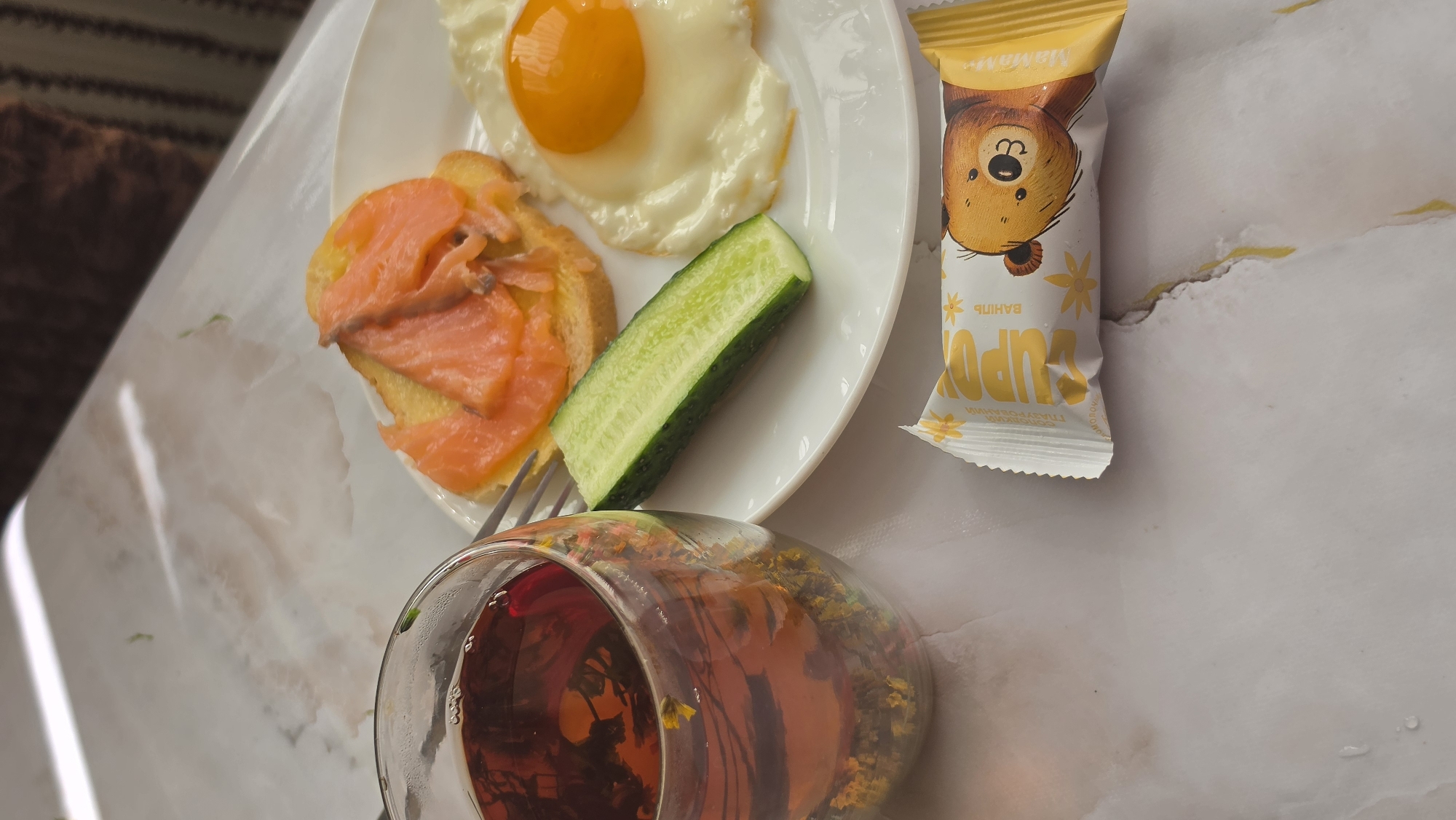In an indoor setting, possibly a kitchen or dining area, a close-up, eye-level shot captures a breakfast spread on a marble-patterned surface. To the left, a clear glass mug filled with dark red tea, containing loose tea leaves and flower petals, sits next to a white plate. On the plate, a slice of bread is topped with vibrant pink smoked salmon. Beside the bread, a piece of green cucumber is laid out. To the right of the cucumber, a fried egg with a bright orange yolk is presented. In the foreground, a wrapper for a snack bar with a cartoon bear illustration and yellow text is visible. The lighting is soft and diffused, creating a warm ambiance. The background is blurred, with hints of brown and white fabric, suggesting furniture or upholstery. There are no people or animals present in the image, and no specific city or location is discernible.